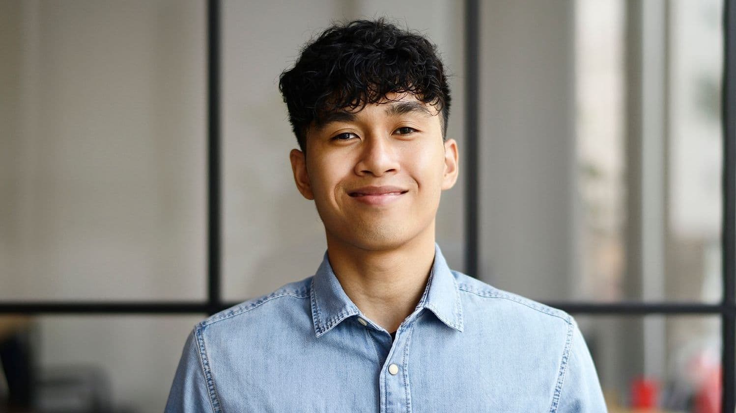 Portrait von einem jungen Mann, der in einem modernen und offenen Büro steht und in die Kamera lächelt.