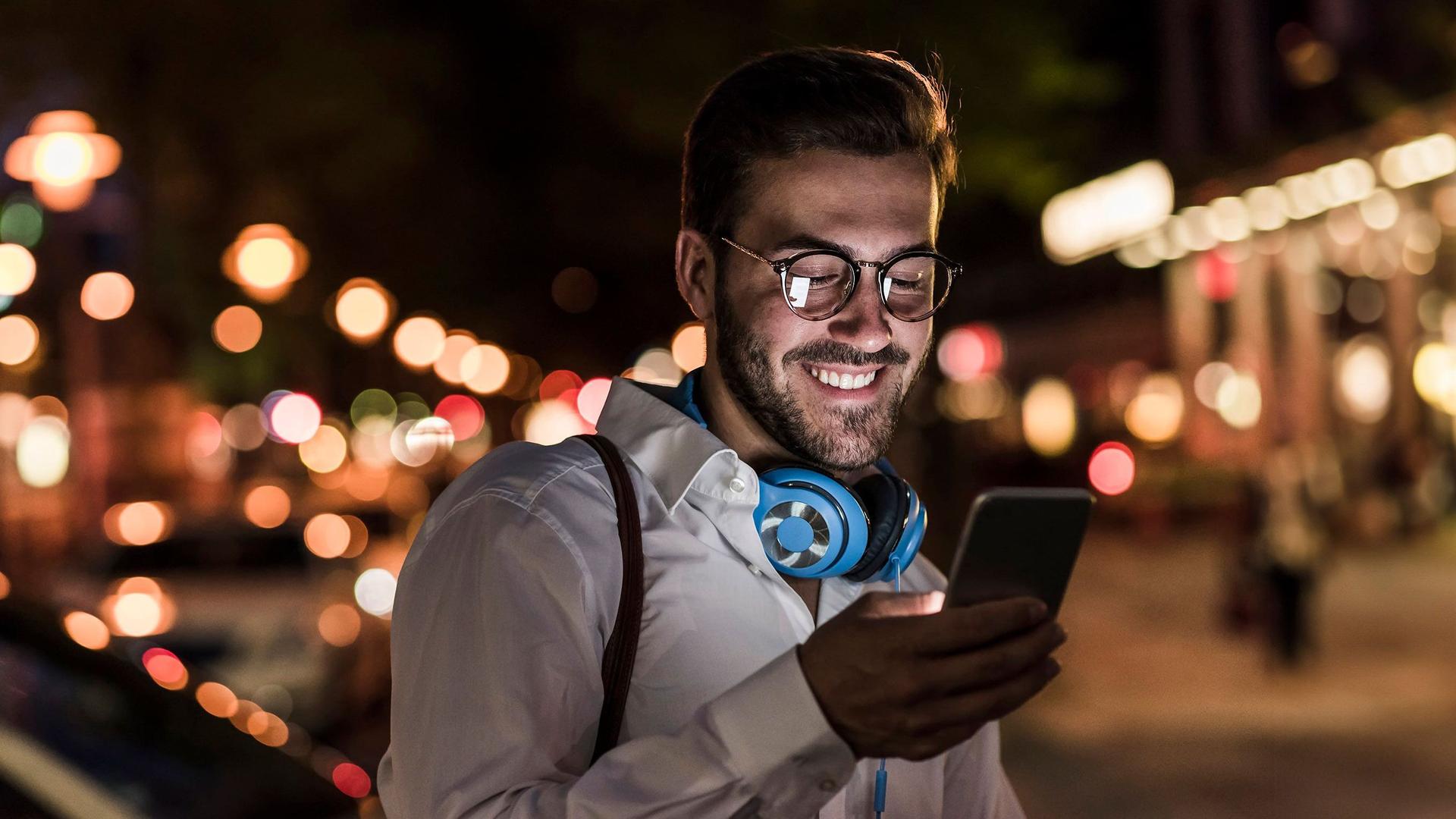 Ein junger Mann im weißen Hemd und türkisen Kopfhörern um den Hals, schaut glücklich auf den Display seines Smartphones. Es ist Abend und sein Gesicht wird vom Displaylicht angeleuchtet.