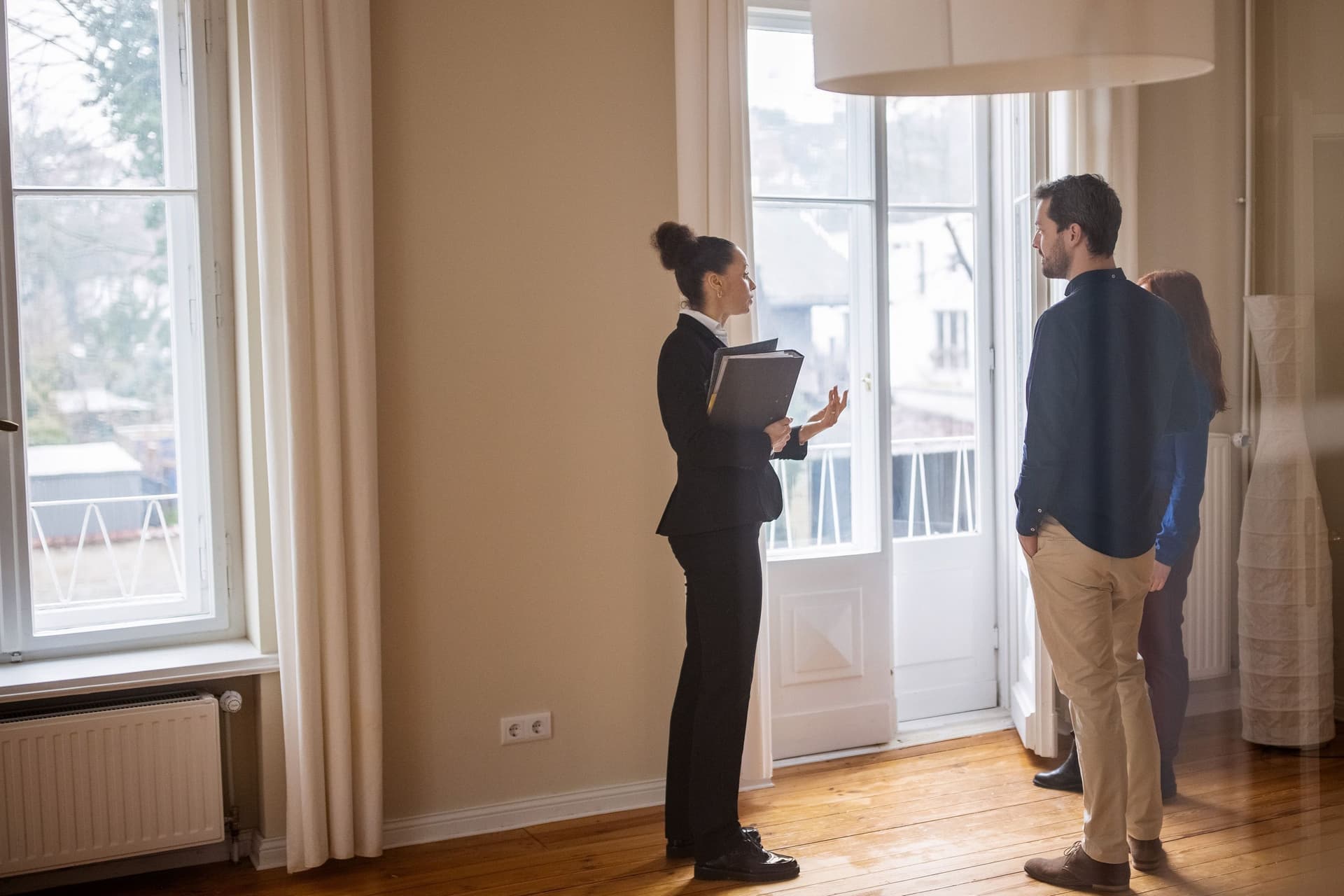 Immobilienmaklerin steht mit einem jungen Paar in einem leeren Zimmer. Sie trägt einen Hosenanzug und hat eine Mappe in der Hand.