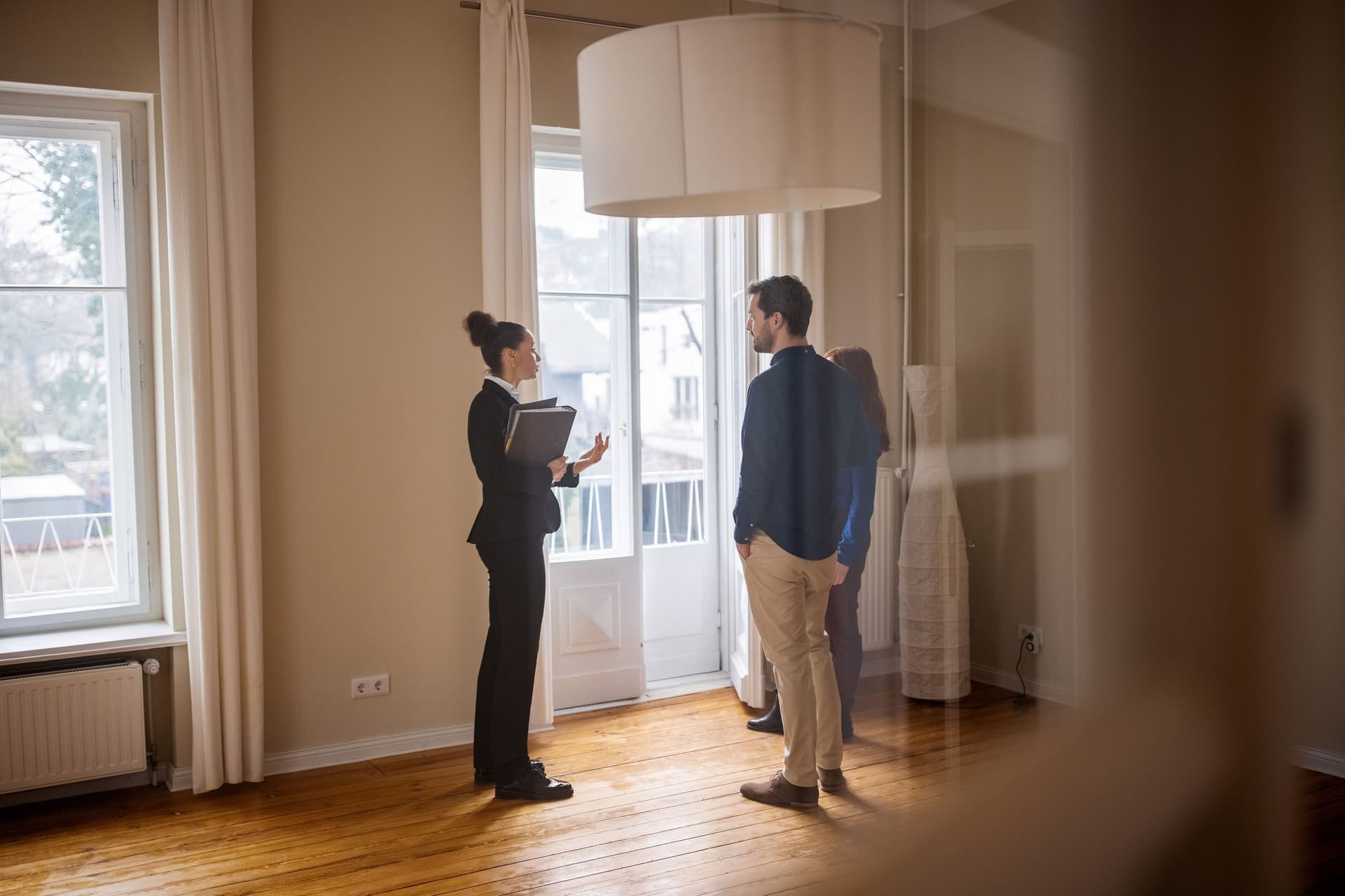 Immobilienmaklerin steht mit einem jungen Paar in einem leeren Zimmer. Sie trägt einen Hosenanzug und hat eine Mappe in der Hand.
