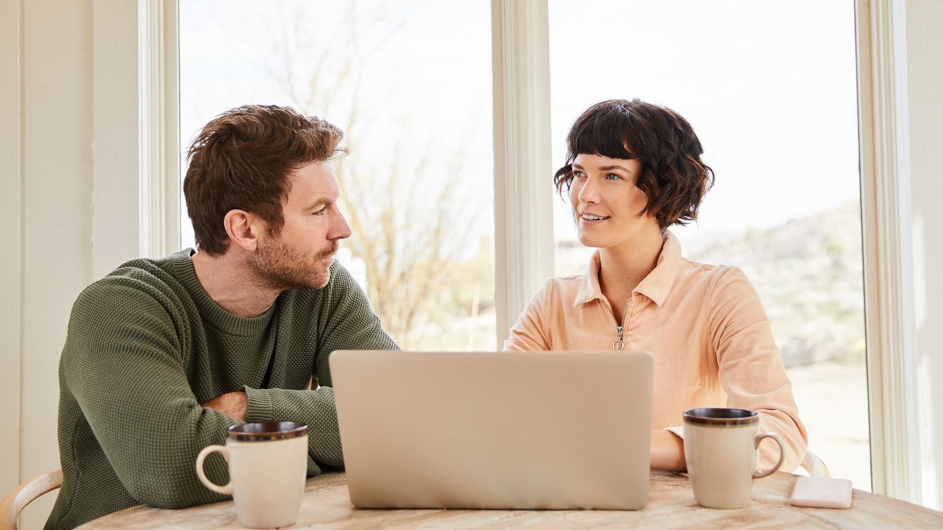 Ein Mann und eine Frau sitzen an einem Tisch vor einem Laptop und unterhalten sich. Beide haben Kaffeetassen vor sich, und durch das große Fenster im Hintergrund fällt Tageslicht in den Raum.