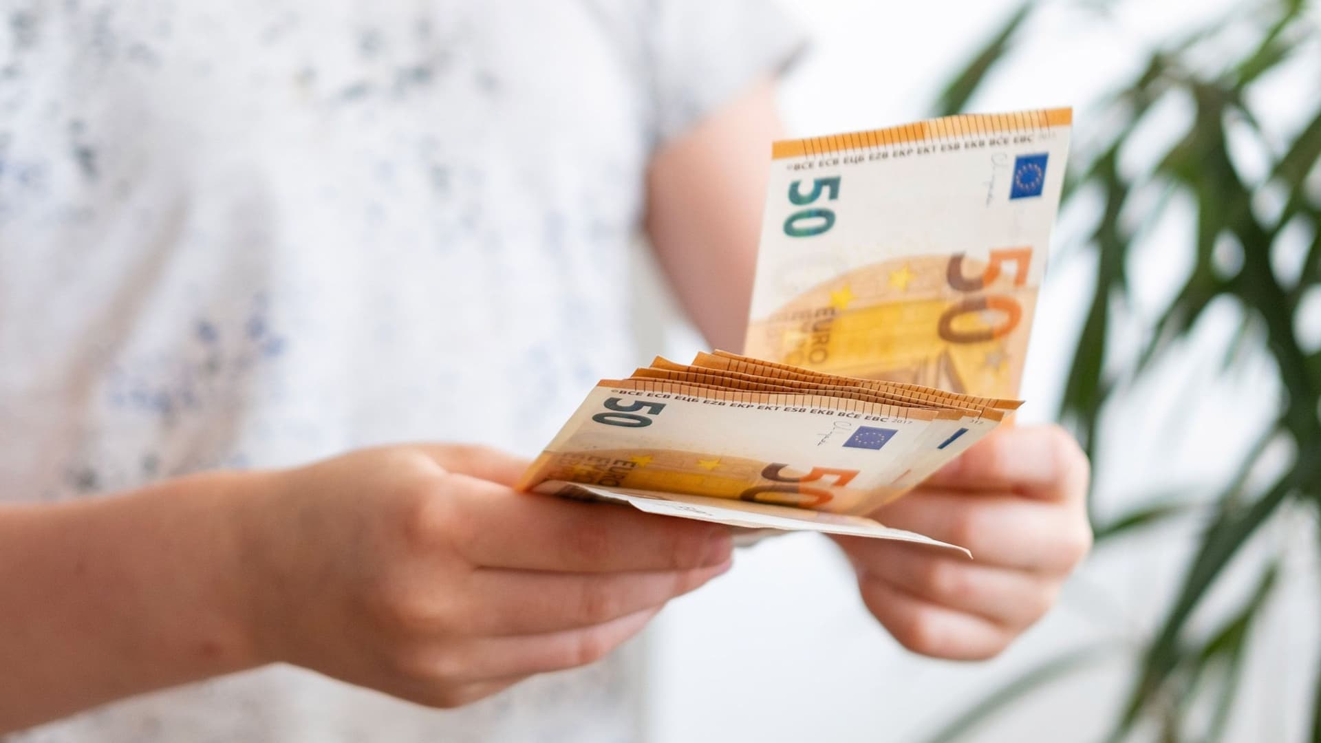 Ein Kind hält ein Bündel mit mehreren Geldscheinen in der Hand. Sie trägt ein helles T-Shirt und eine Pflanze ist im Hintergrund erkennbar.