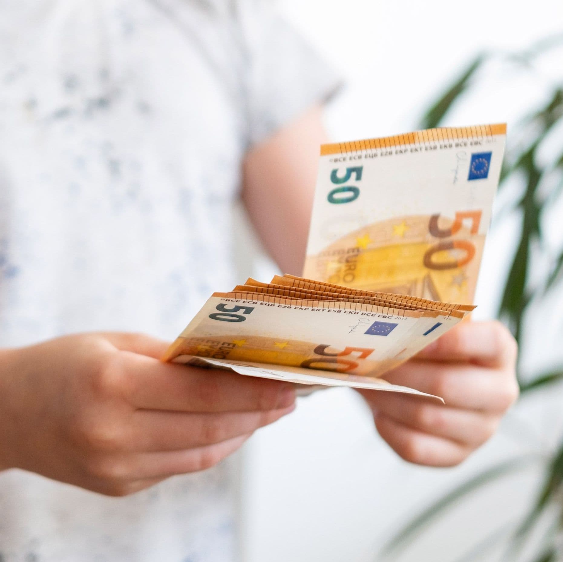 Ein Kind hält ein Bündel mit mehreren Geldscheinen in der Hand. Sie trägt ein helles T-Shirt und eine Pflanze ist im Hintergrund erkennbar.