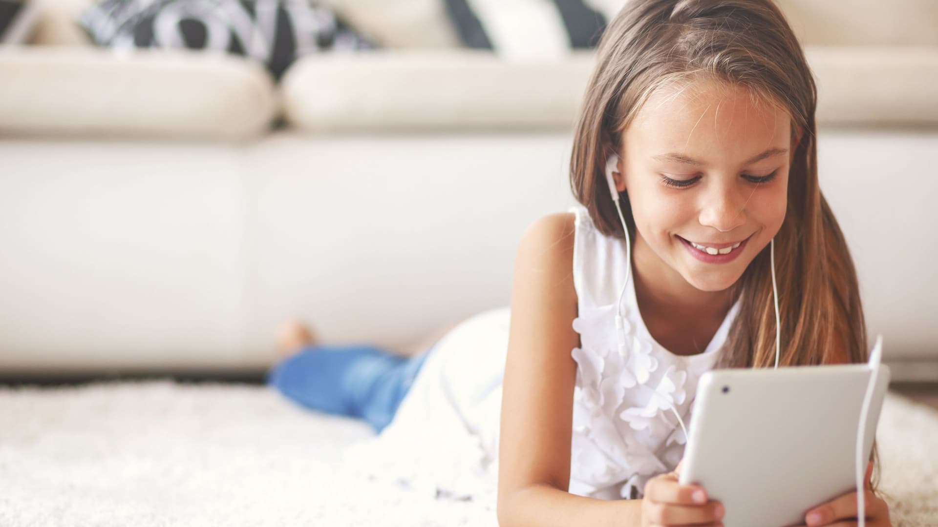 Mädchen im Grundschulalter liegt auf weißem Teppich mit Tablet in den Händen. Sie lächelt und hat Kopfhörer im Ohr.