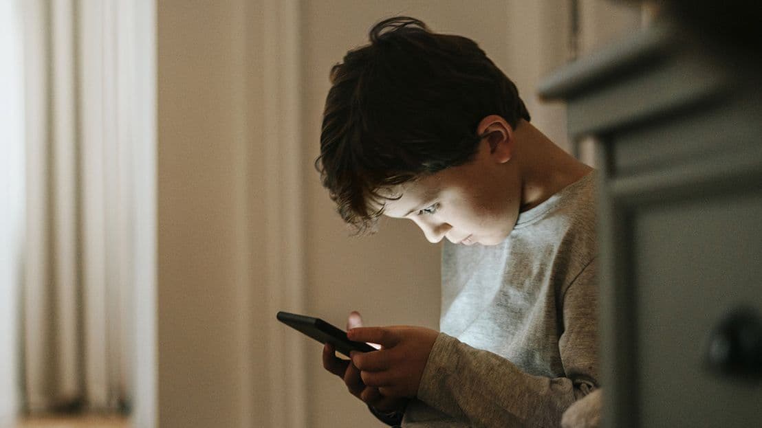 Ein Junge sitzt in einem schwach beleuchteten Raum und blickt konzentriert auf ein Smartphone, dessen Bildschirm sein Gesicht erhellt.