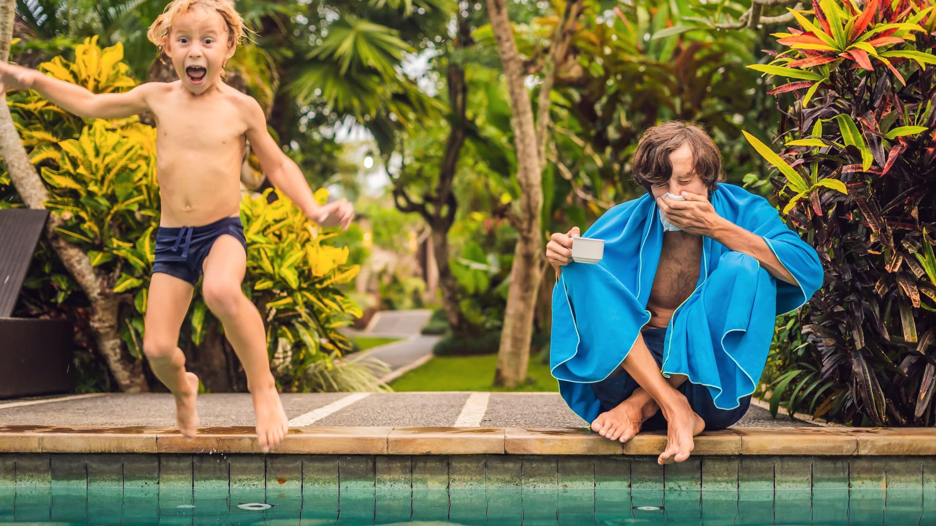 Verschnupfter Vater mit Kind am Pool. Das Kind springt lachend in den Pool während sich der Vater die Nase putzt.