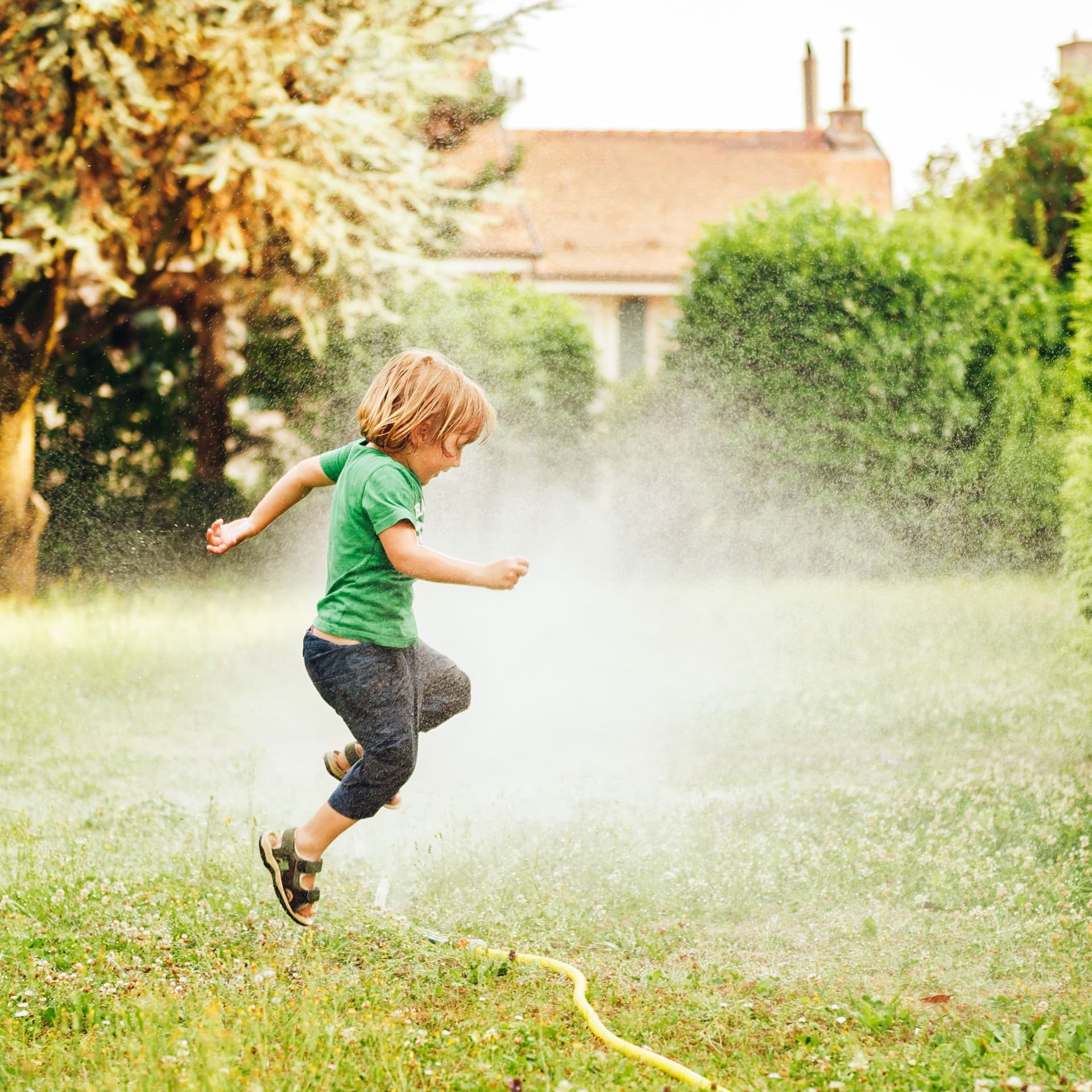 Kind springt über einen Gartensprinkler auf einer Wiese.
