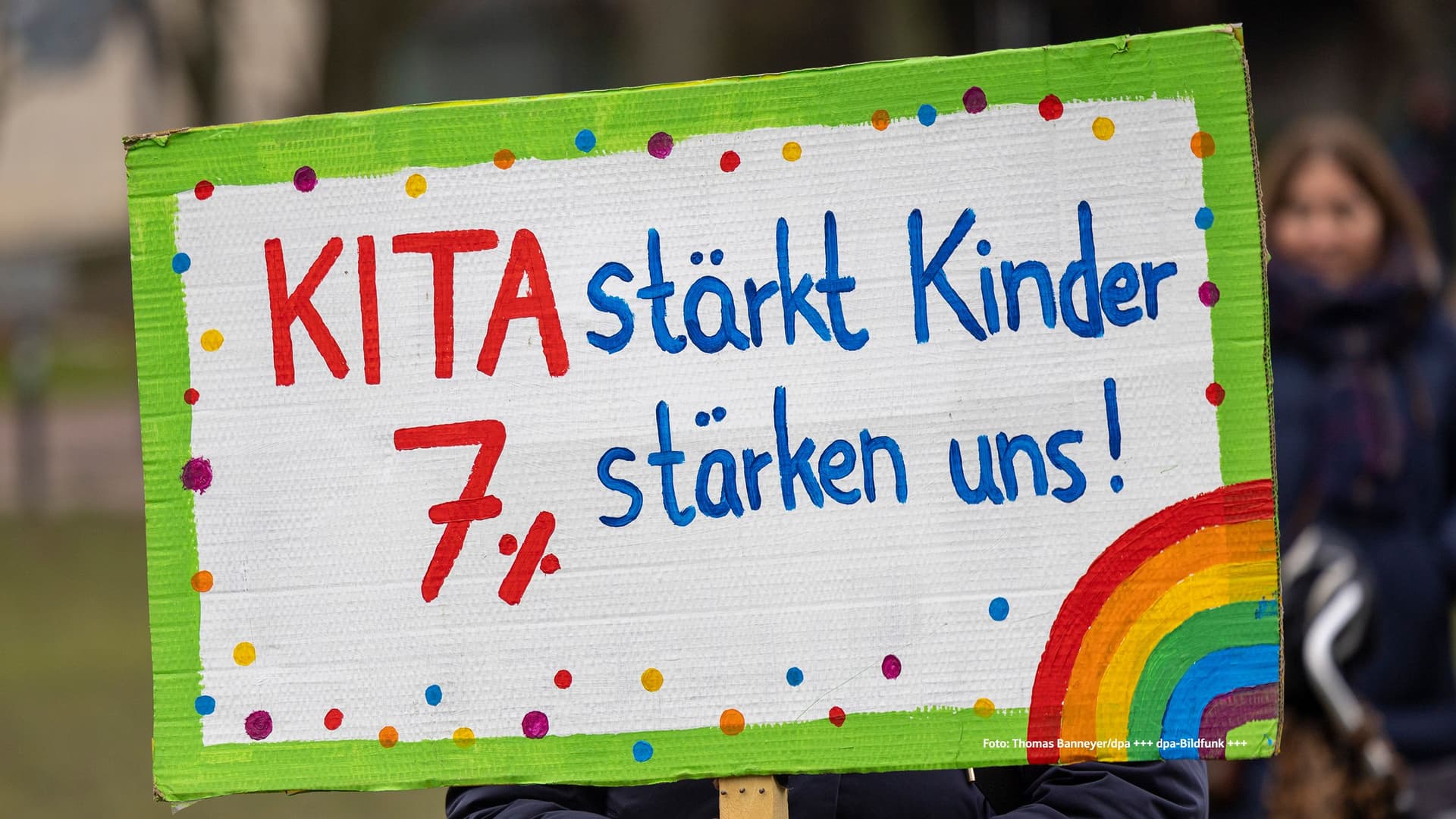 Handgemaltes Protestschild mit grünem Rand und Regenbogen, darauf der Text: „KITA stärkt Kinder 7% stärken uns!“ — Fotografie eines Demonstrationsplakats, gehalten von einer Person im Hintergrund.