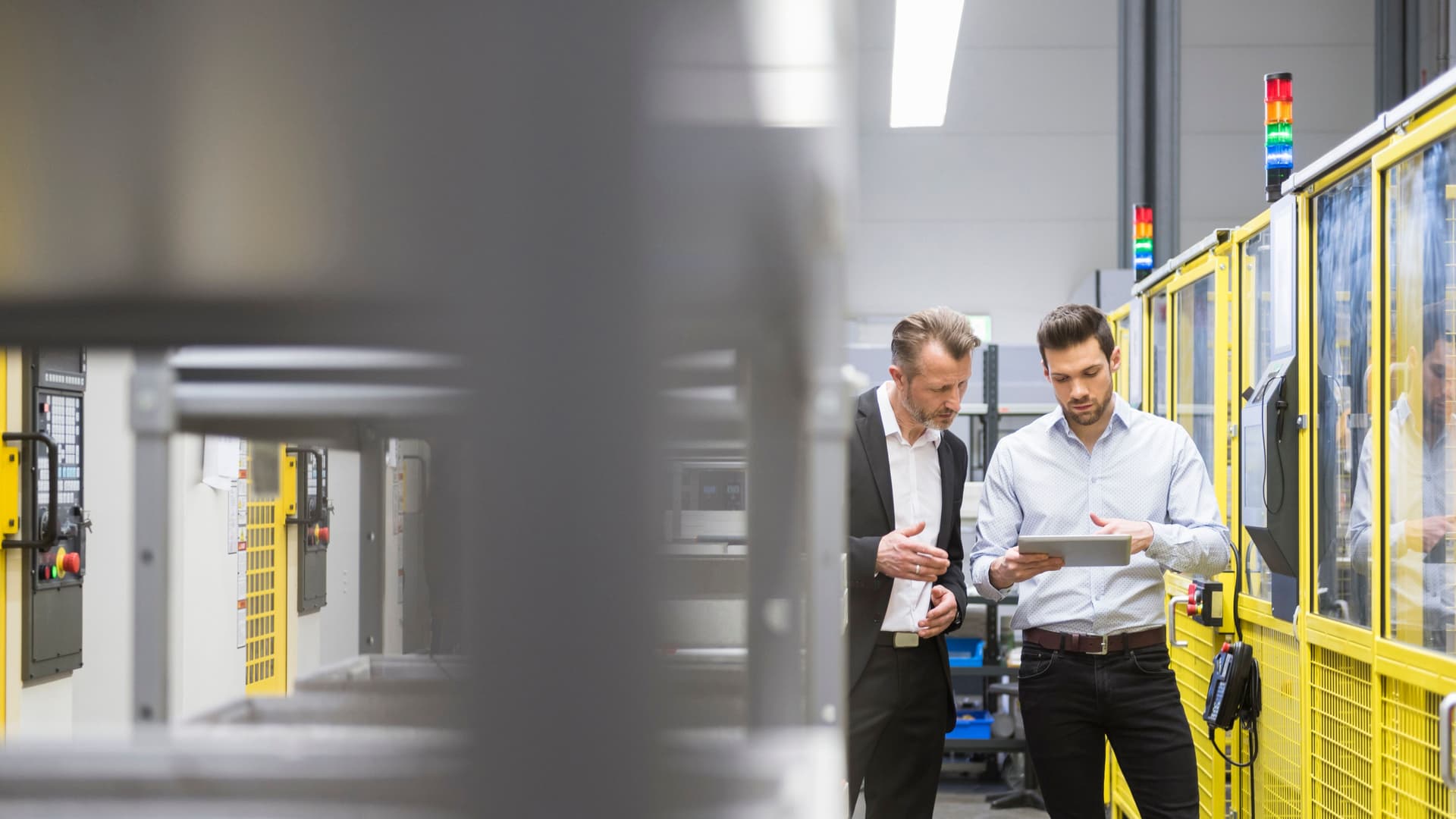 Zwei Männer in Anzug stehen in einer Fabrik mit Industrierobotern  und gucken zusammen auf ein Tablet.