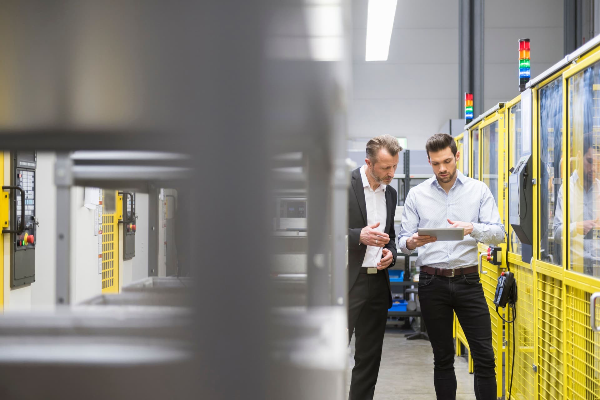 Zwei Männer in Anzug stehen in einer Fabrik mit Industrierobotern  und gucken zusammen auf ein Tablet.