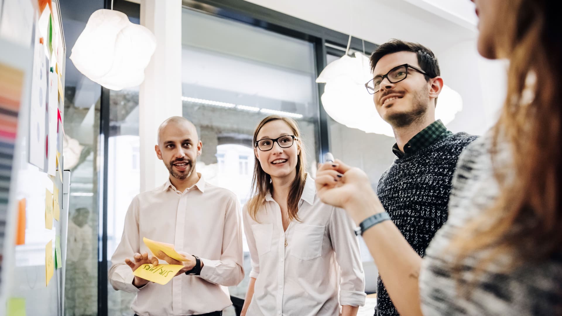 Ein Team junger Leute bespricht in einem modernen Büro ihr Projekt. Die Gruppe besteht aus zwei Männern und zwei Frauen. Sie stehen vor einer Tafel mit Notizen und Diagrammen.
