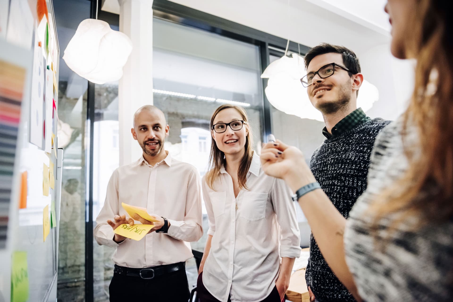 Ein Team junger Leute bespricht in einem modernen Büro ihr Projekt. Die Gruppe besteht aus zwei Männern und zwei Frauen. Sie stehen vor einer Tafel mit Notizen und Diagrammen.