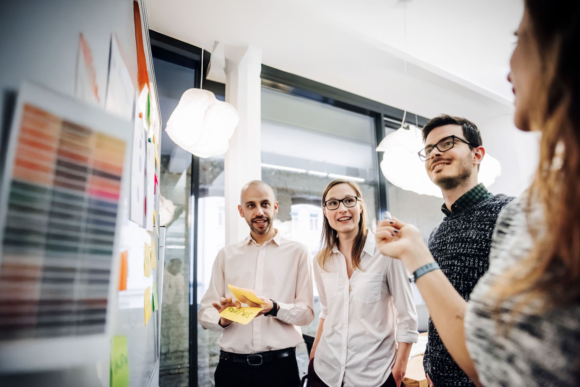 Ein Team junger Leute bespricht in einem modernen Büro ihr Projekt. Die Gruppe besteht aus zwei Männern und zwei Frauen. Sie stehen vor einer Tafel mit Notizen und Diagrammen.