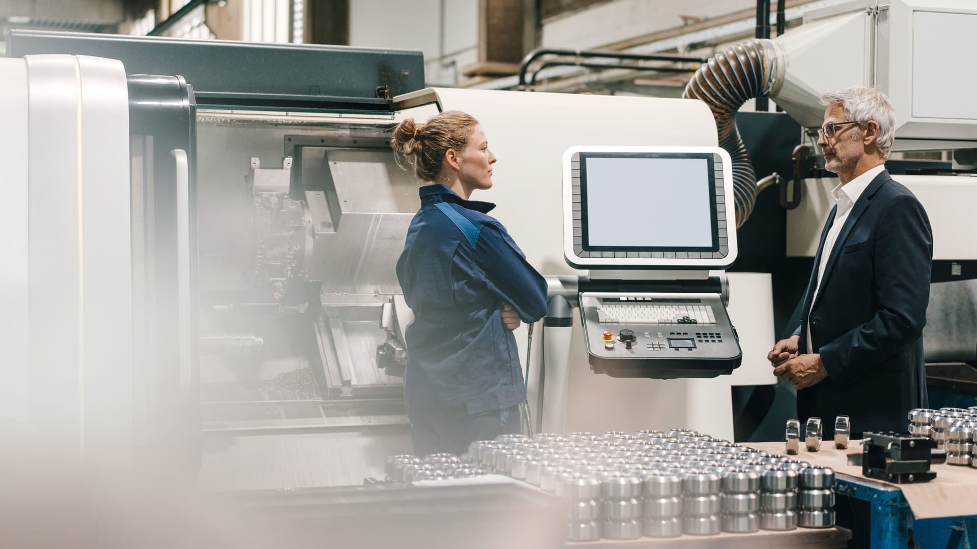 Frau und Mann stehen vor einer Maschine mit Bildschirm in einer Produktionshalle.