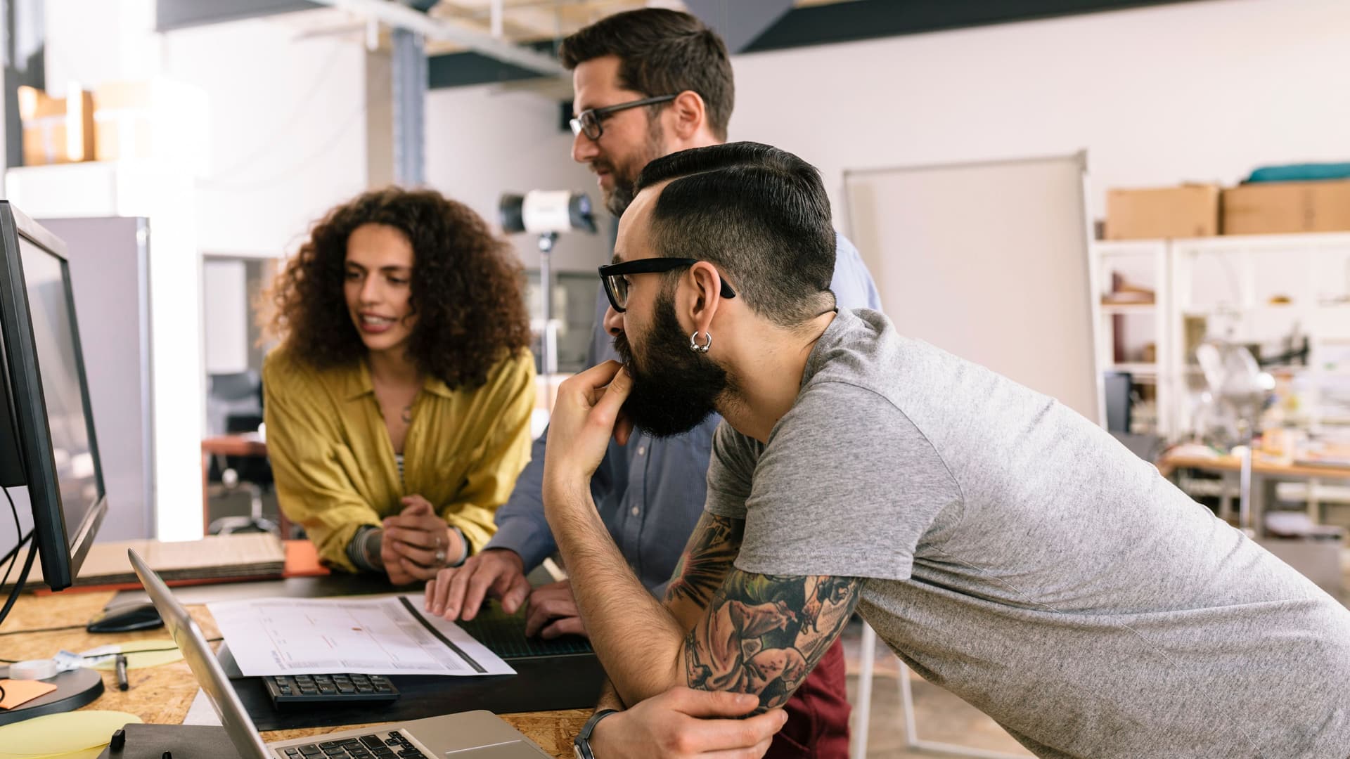 Zwei Männer und eine Frau arbeiten gemeinsam stehend vor einem PC in einem Startup-Büro.
