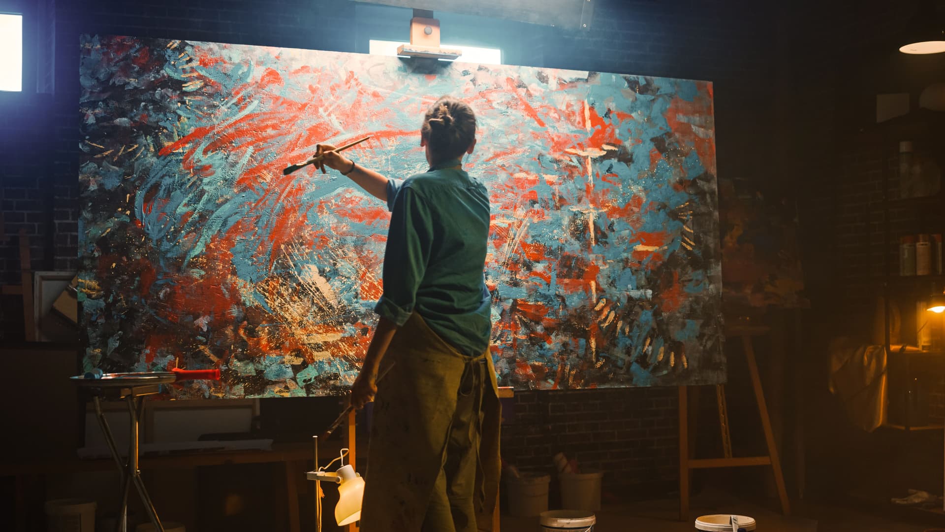 Person mit Pinsel in der Hand von hinten, die eine Leinwand mit Farbe bemalt. Licht-Spotlight ist auf das Bild gerichtet.
