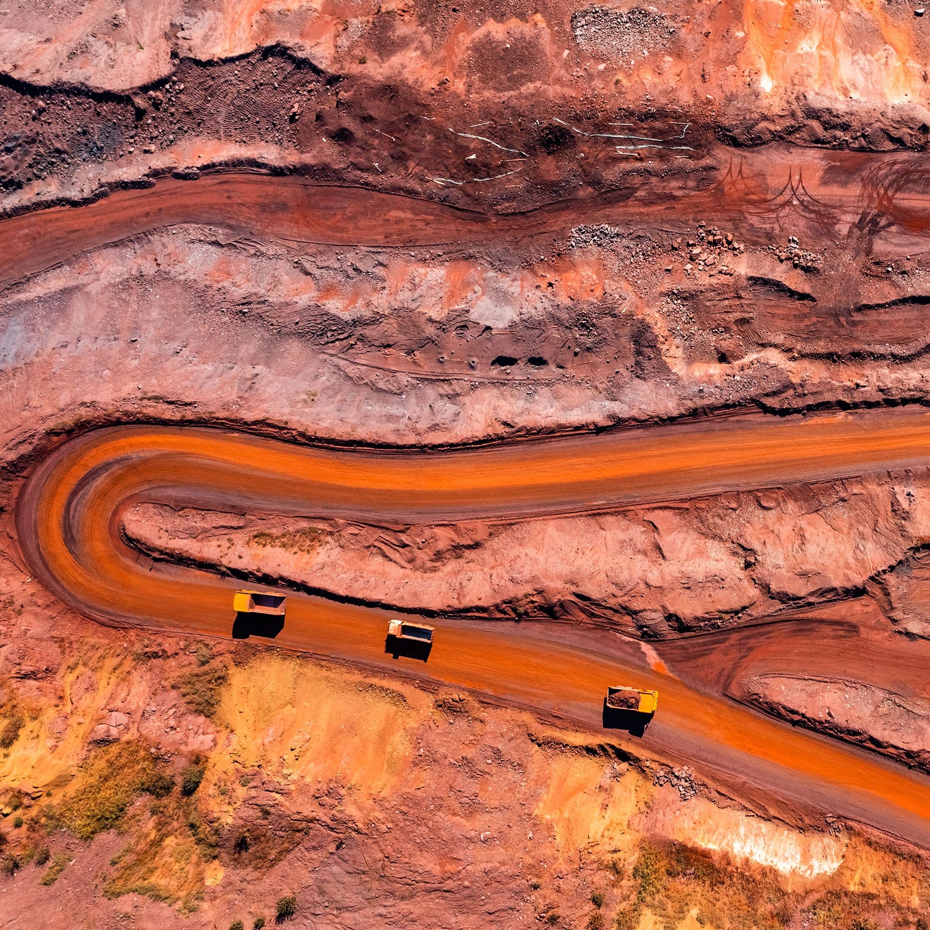 Luftaufnahme einer Kupfermine mit geschwungenen rostbraunen Wegen und mehreren gelben Muldenkippern, die Material transportieren; die Umgebung zeigt abgestufte Abraumflächen und rötlich-orangefarbenen Boden.