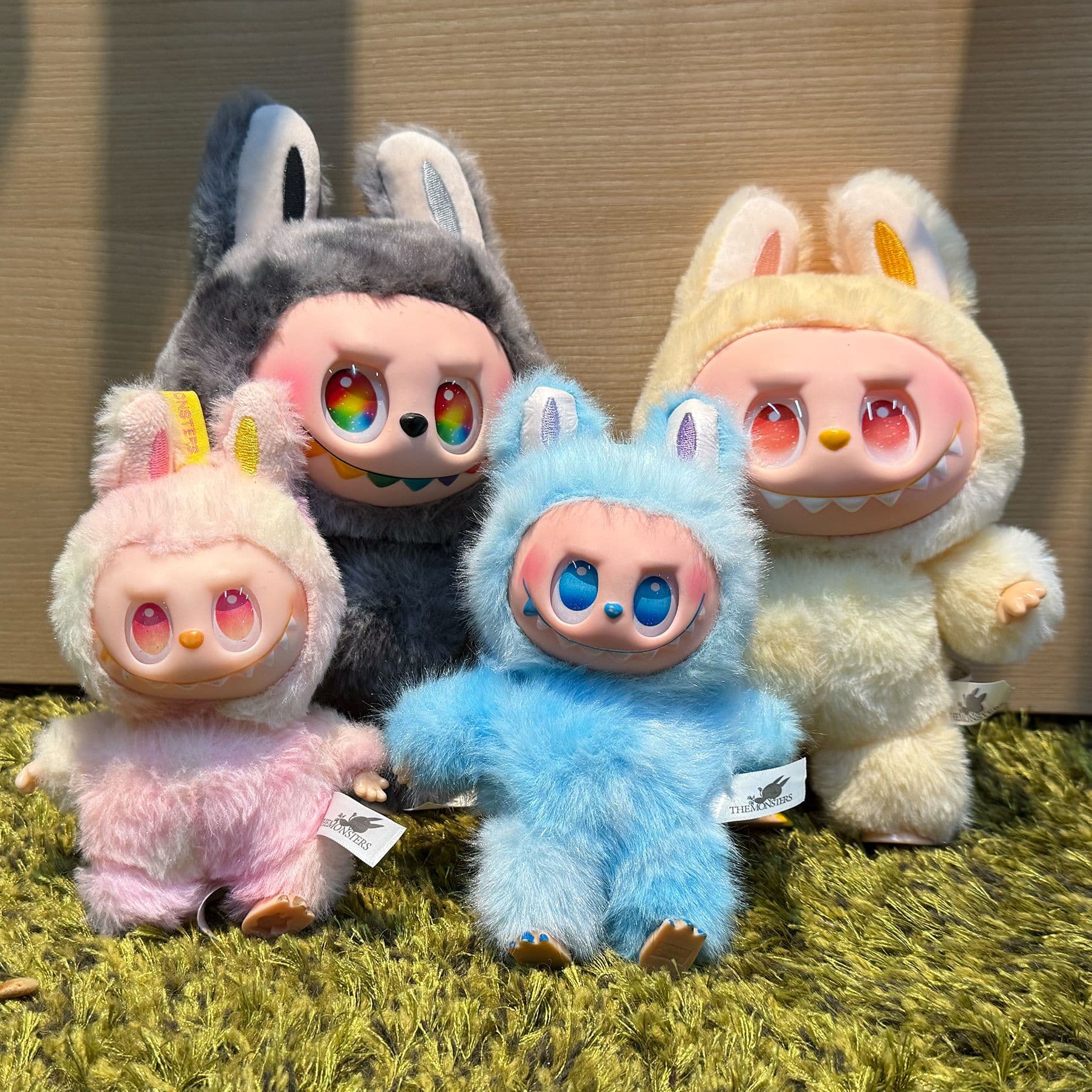 Vier bunte Sammelfiguren in flauschigen Tierkostümen (Labubus), die nebeneinander auf einem grünen Teppich sitzen. 