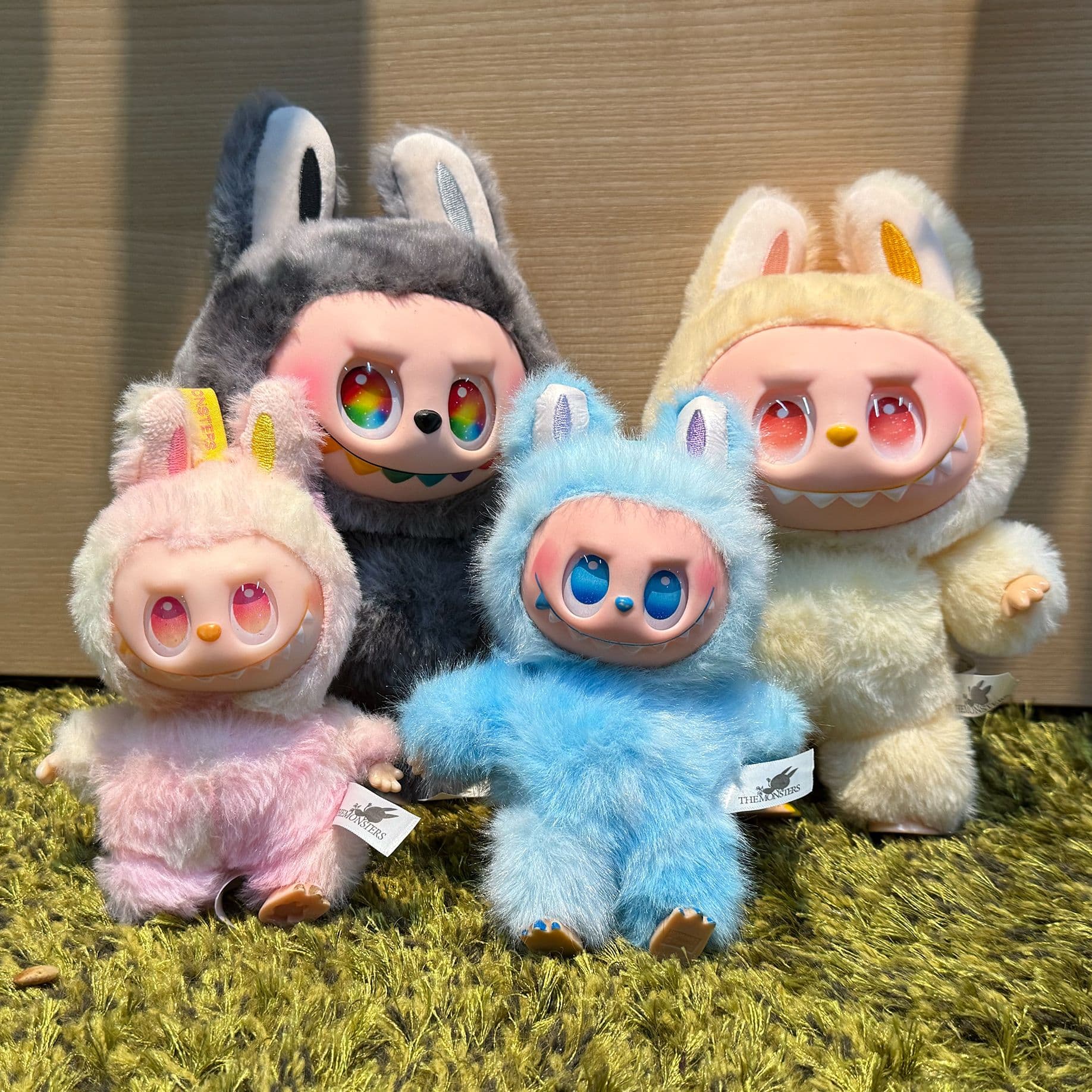 Vier bunte Sammelfiguren in flauschigen Tierkostümen (Labubus), die nebeneinander auf einem grünen Teppich sitzen. 
