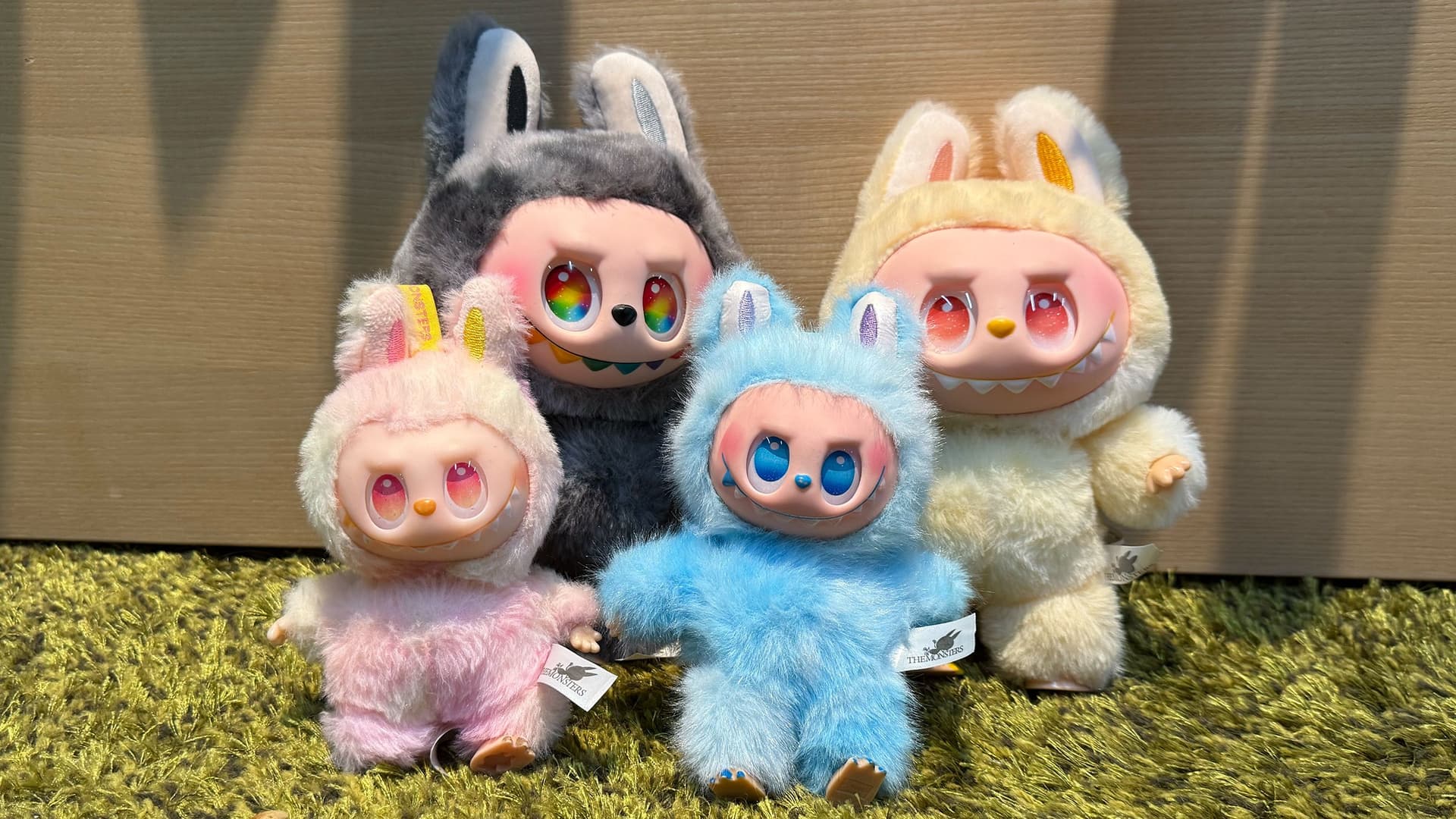 Vier bunte Sammelfiguren in flauschigen Tierkostümen (Labubus), die nebeneinander auf einem grünen Teppich sitzen. 