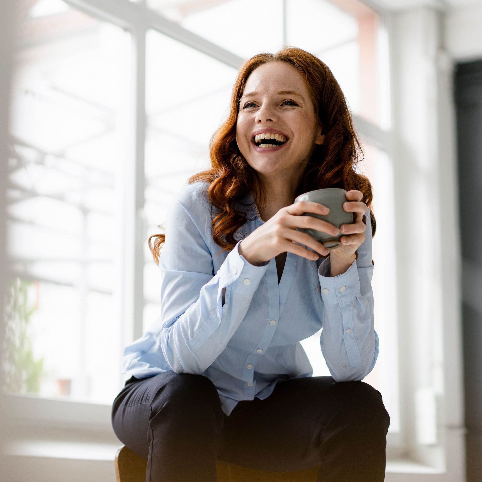 Portrait of laughing redheaded woman with tea bowl sitting on backrest in a loft
