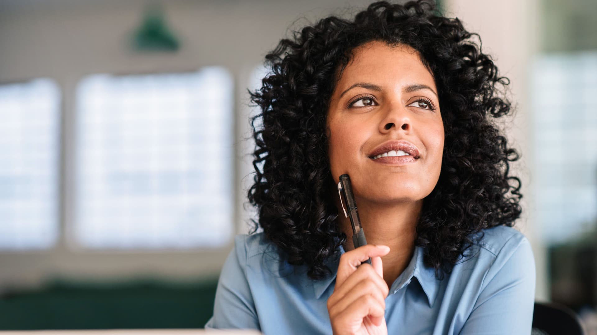 Lächelnde junge Frau guckt tief in Gedanken nach oben und hält einen Stift in der Hand. Sie sitzt an einem Schreibtisch in einem Büro mit vielen Fenstern.