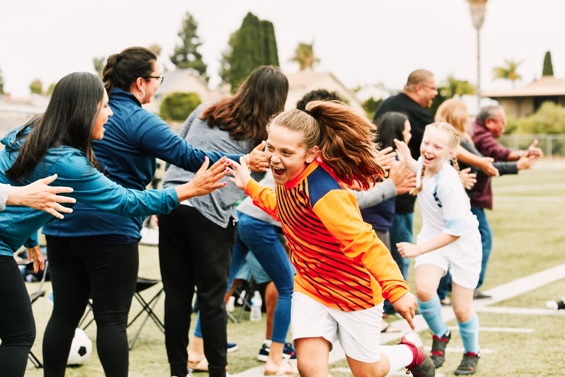 Mädchen in Fußballtrikots klatschen jubelnd auf einem Fußballplatz mit den am Rande stehenden Frauen und Männern ab.