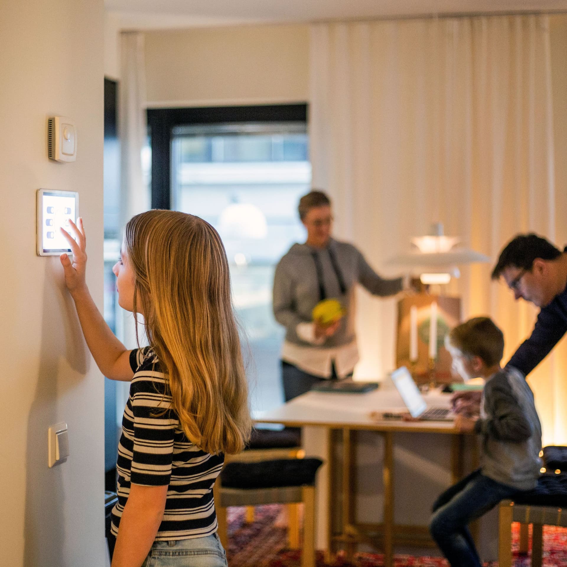 Mädchen bedient digitales Smart Home Display an der Wand. Im Hintergrund ist unscharf die Familie zu erkennen.