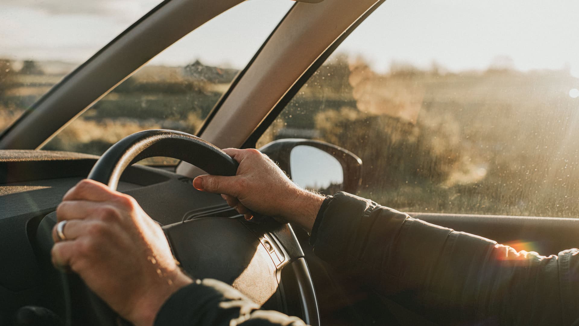 Hände umgreifen ein Autolenkrad. Von der Seite scheint die Sonne in das Auto.