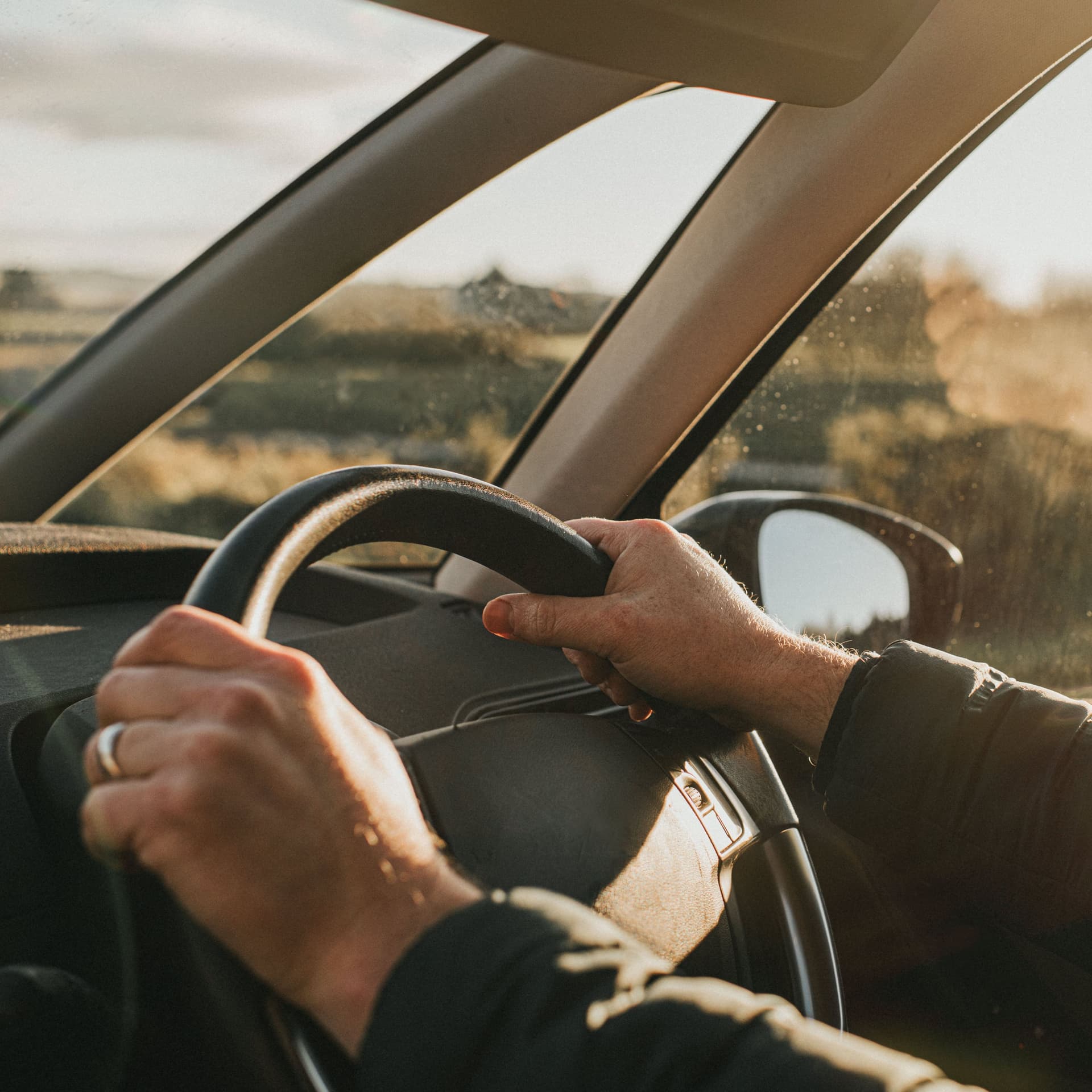 Hände umgreifen ein Autolenkrad. Von der Seite scheint die Sonne in das Auto.