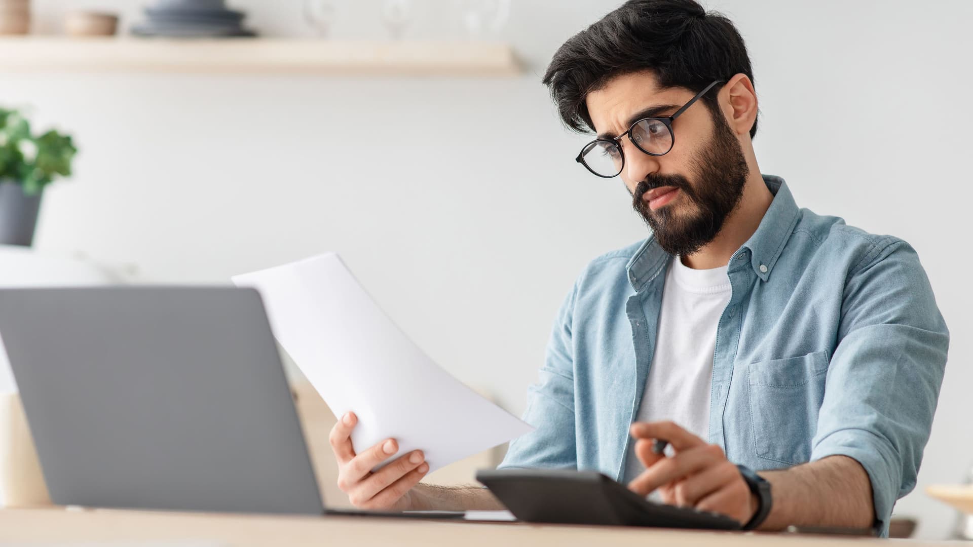 Nicht weißer Mann Steuern berechnet oder Budgetiert am Küchentisch mit Laptop und Papieren.