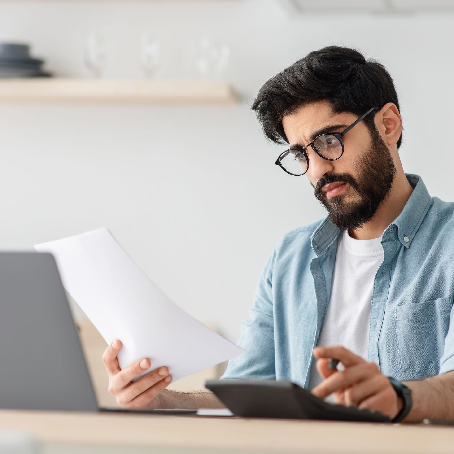 Nicht weißer Mann Steuern berechnet oder Budgetiert am Küchentisch mit Laptop und Papieren.