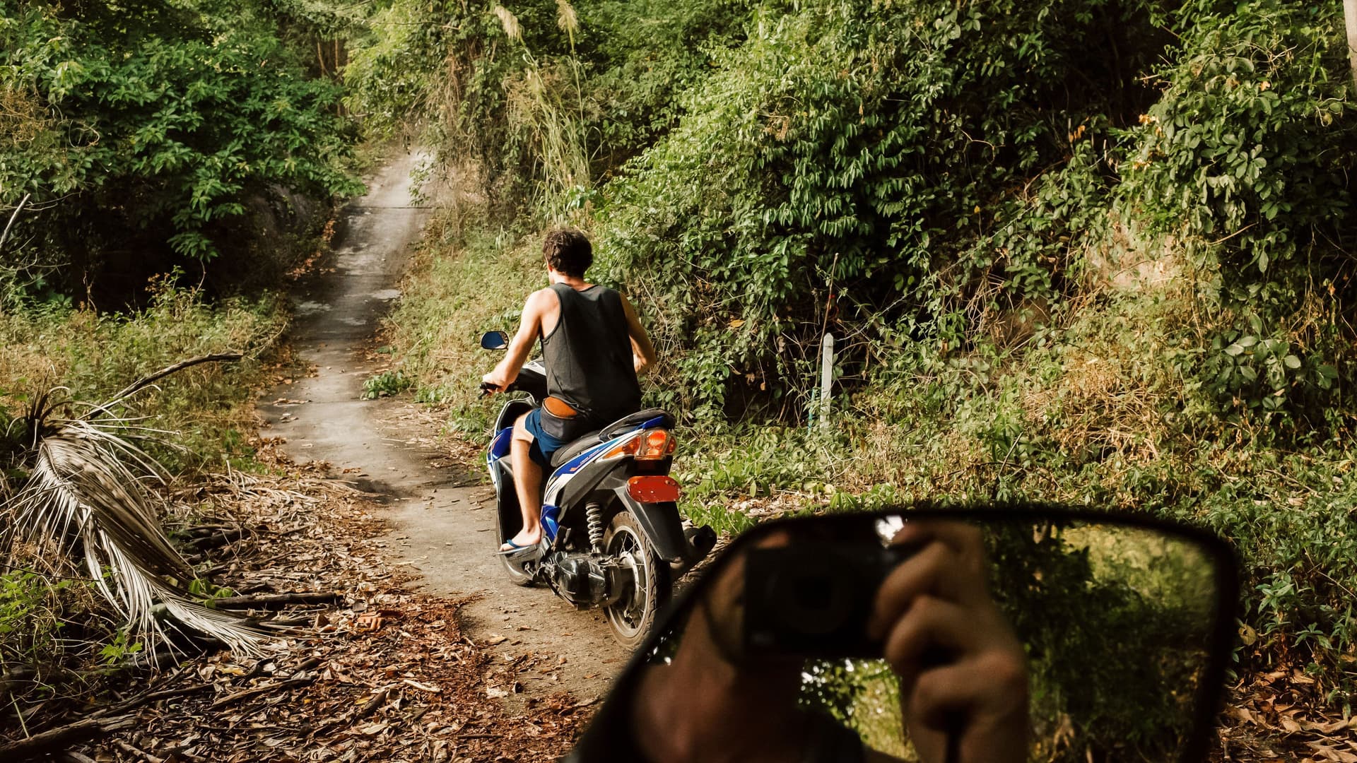 Junger Mann fährt auf Scooter einen Dschungelpfad entlang. Ein anderer Mann spiegelt sich im Rückspiegel während er ein Foto macht.