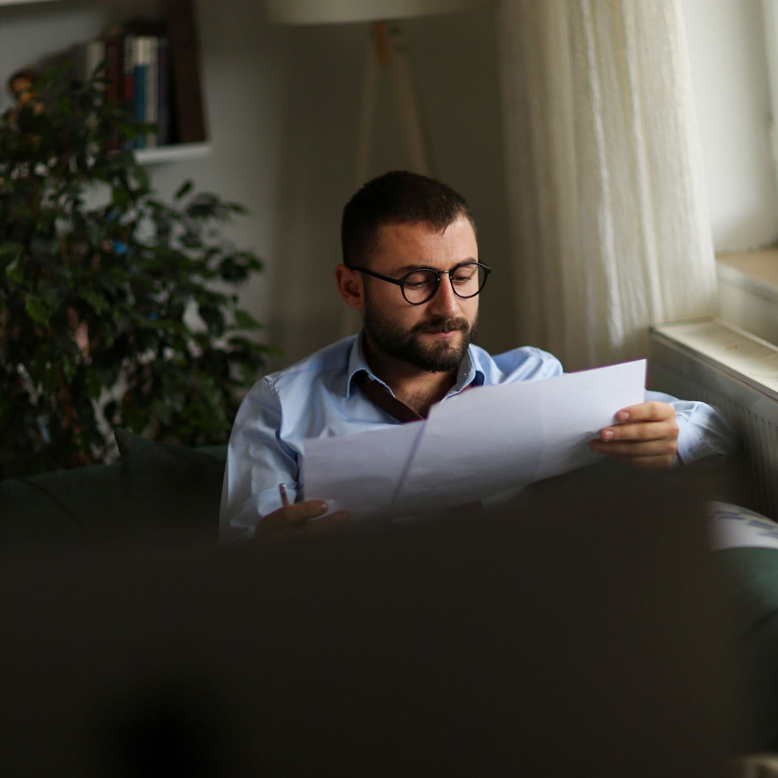 Ein Mann sitzt konzentriert auf einem Sofa und betrachtet aufmerksam ausgedruckte Dokumente. Im Hintergrund stehen eine Zimmerpflanze und ein Bücherregal. 