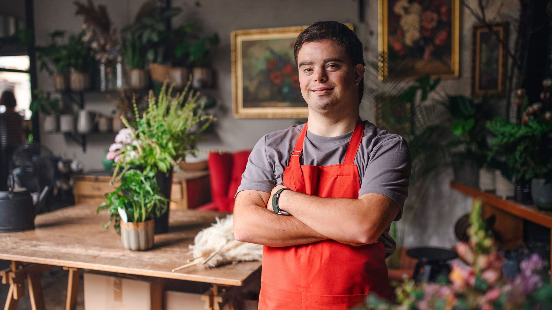 Junger Florist mit Down-Syndrom steht in einem Blumenladen, schaut in die Kamera und lächelt.