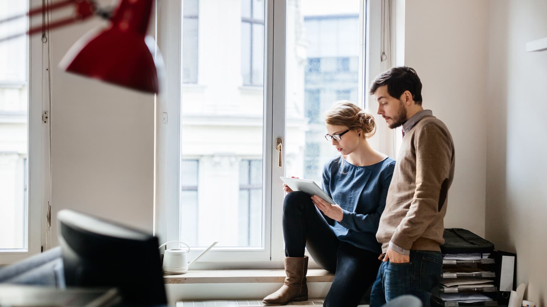 Eine junge Frau sitzt an einem Fenster in einem Büro und neben ihr steht ein junger Mann. Beide schauen auf ein digitales Tablet.