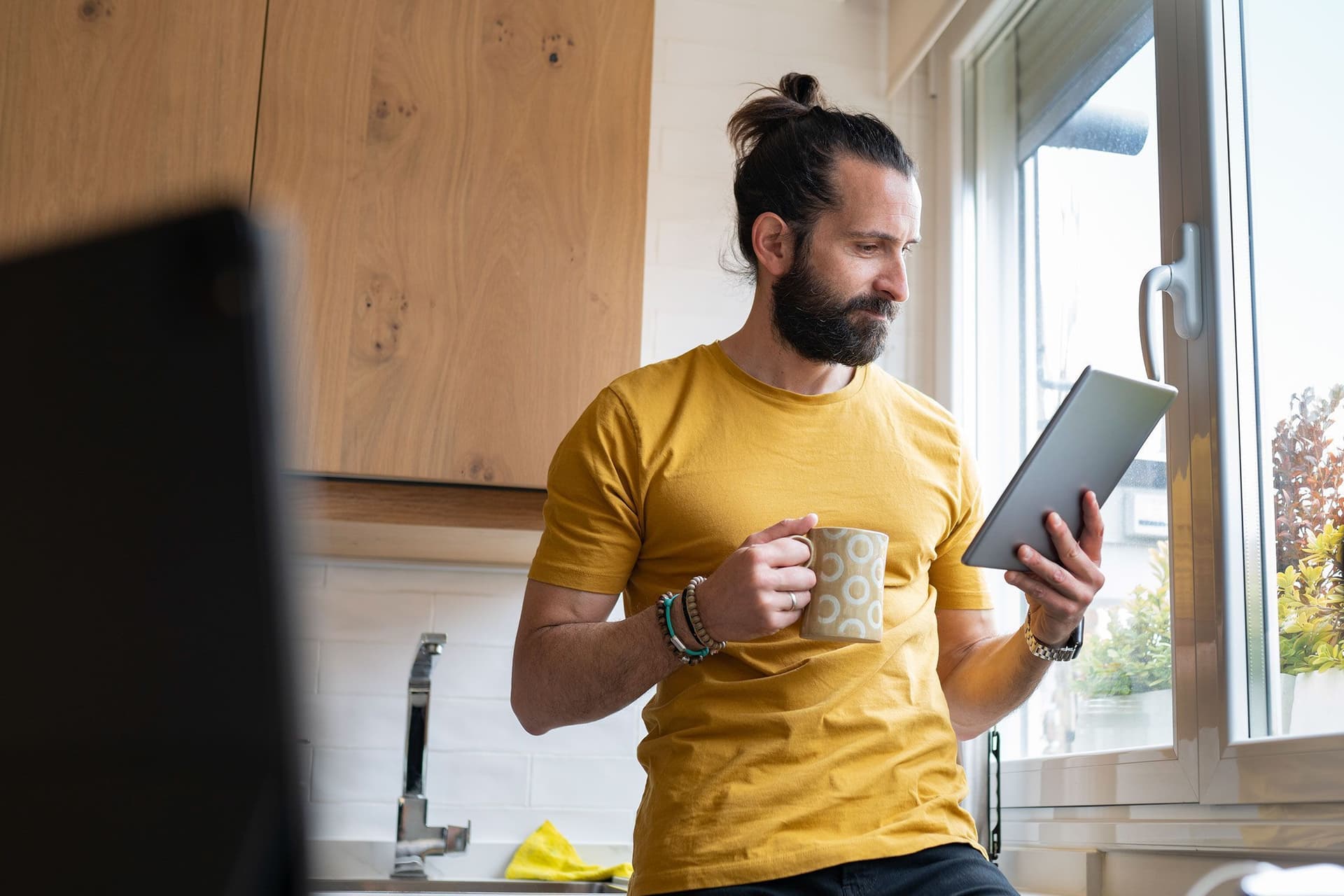 Ein junger Mann lehnt mit einem Kaffeebecher an einer Küchenzeile neben dem Fenster. Er schaut konzentriert auf ein Tablet in seiner Hand.