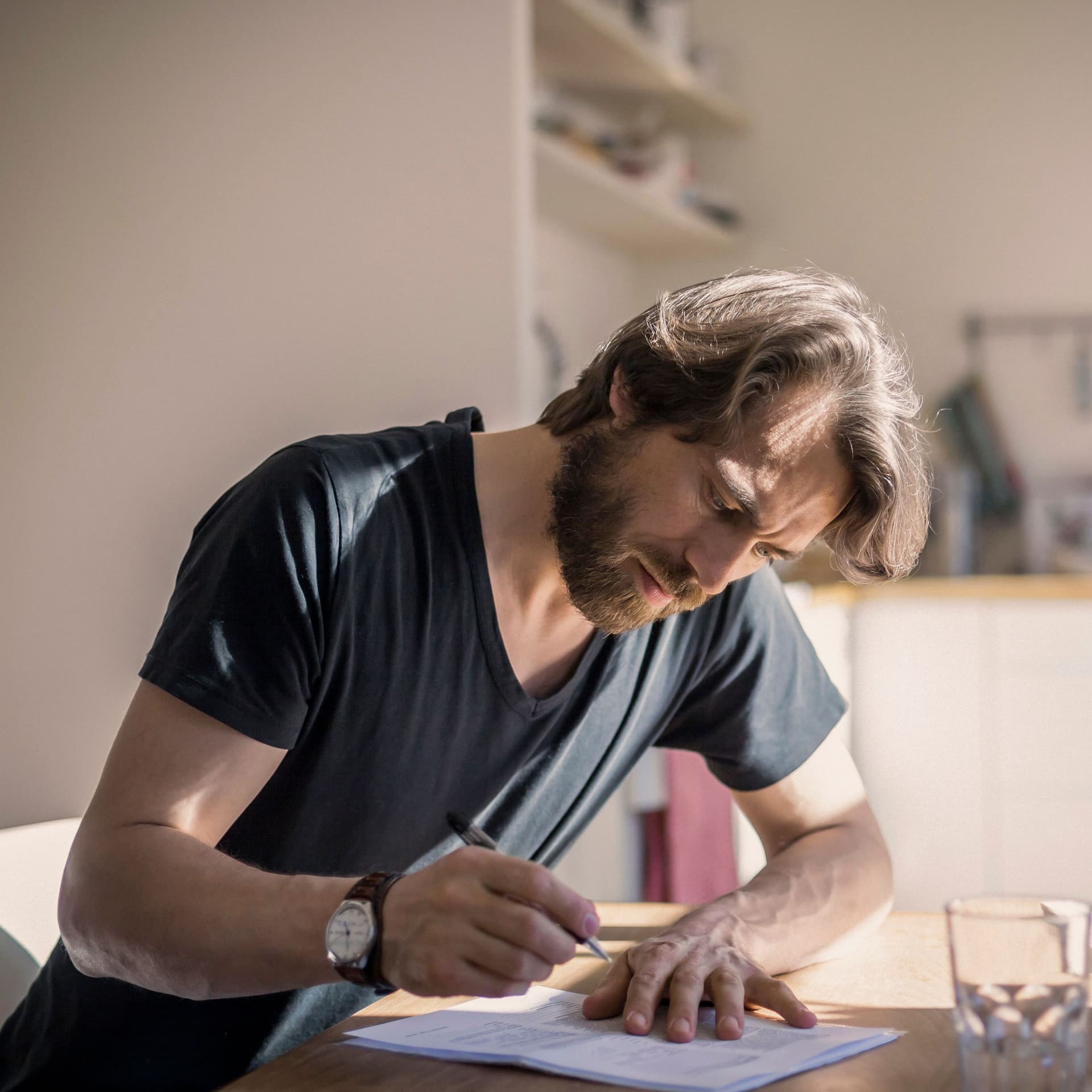 Bärtiger Mann unterzeichnet ein Papier am Küchentisch. Sonnenlicht fällt in die Küche.