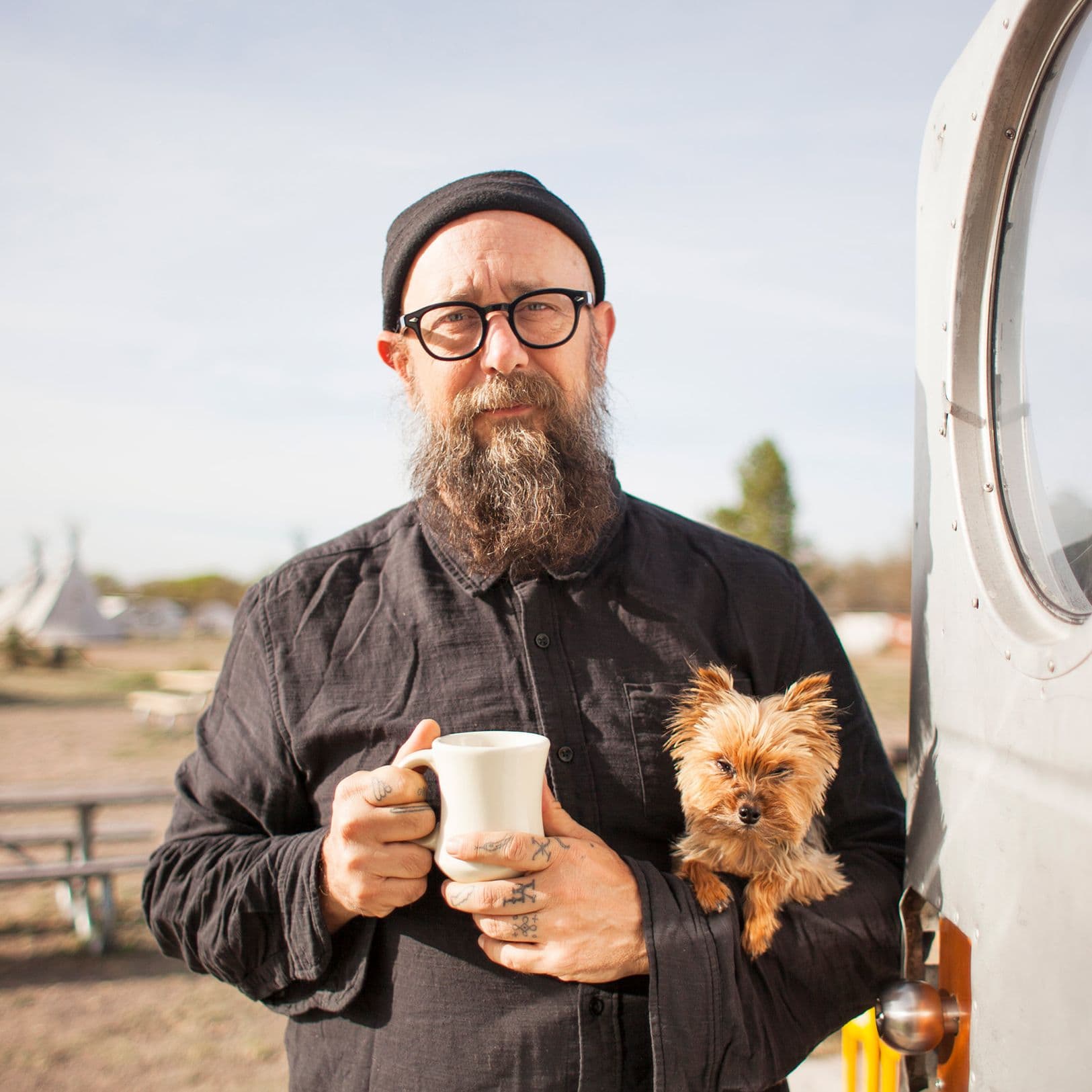 Ein bärtiger Mann mit schwarzer Mütze und Brille steht vor einem Wohnwagen, hält eine weiße Tasse in der Hand und trägt einen kleinen Hund auf dem Arm. Im Hintergrund sind ein Campingplatz und Tipi-Zelte zu sehen.