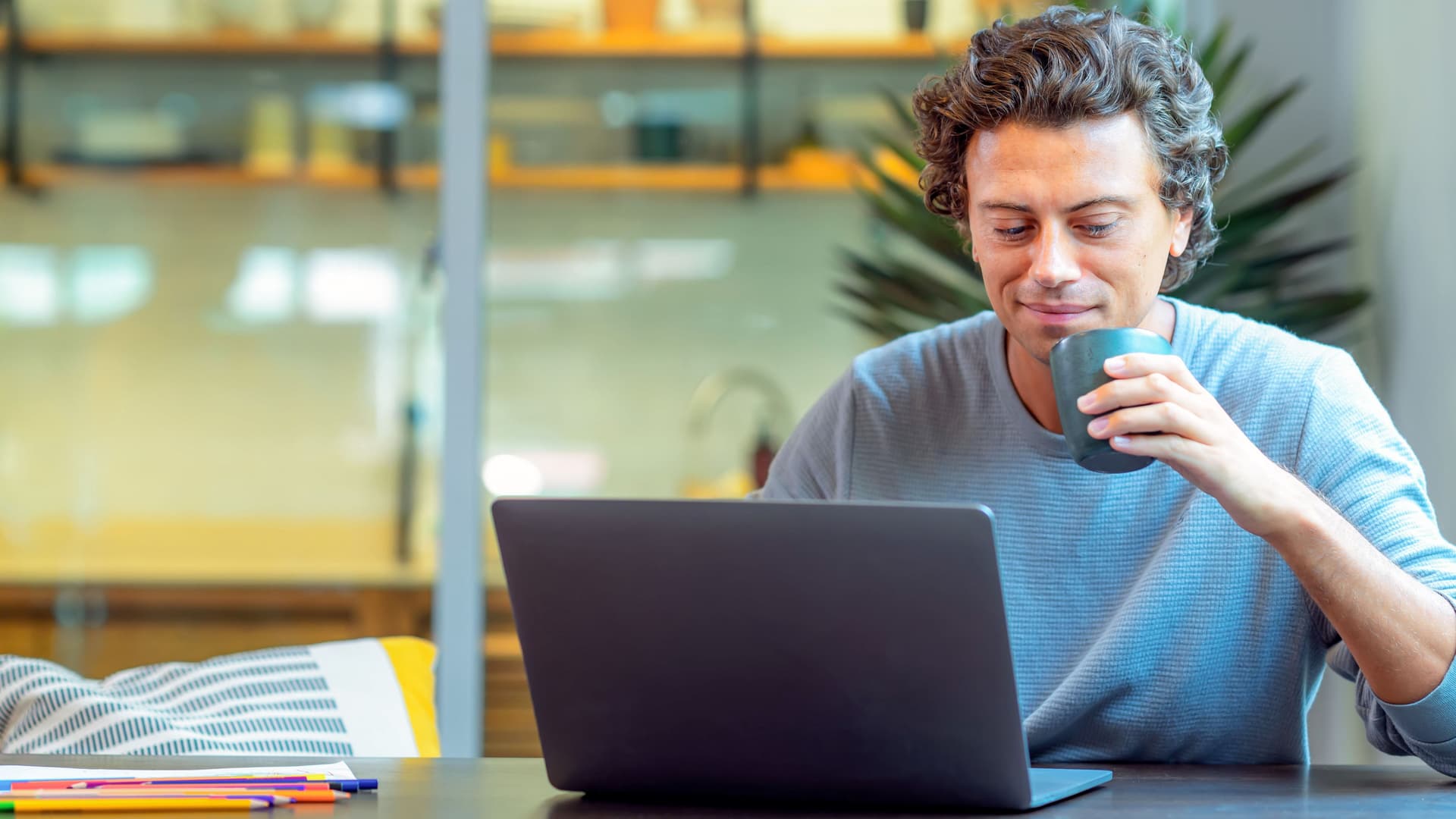 Ein junger Mann sitzt lächelnd und mit Kaffeetasse in der Hand an einem Laptop Computer.