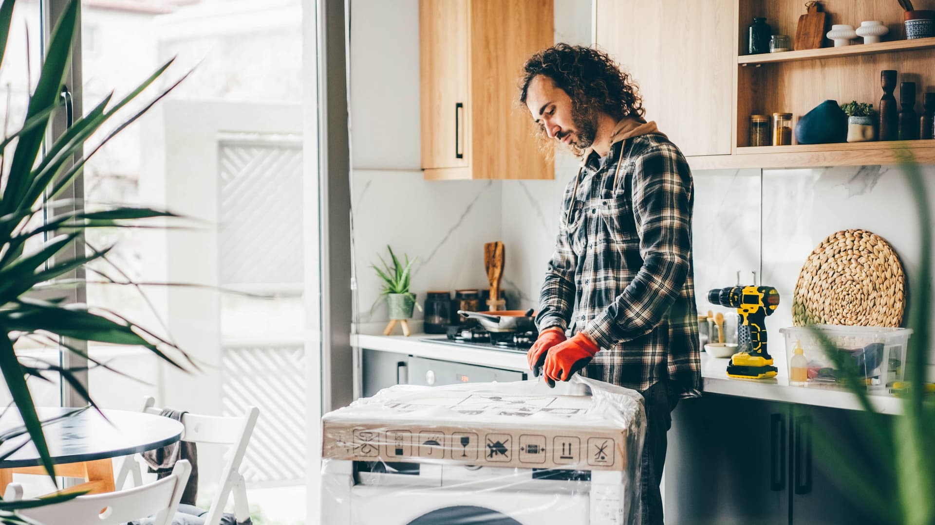 Junger Mann mit roten Arbeitshandschuhen steht in einer Küche mit einer unausgepackten Waschmaschine