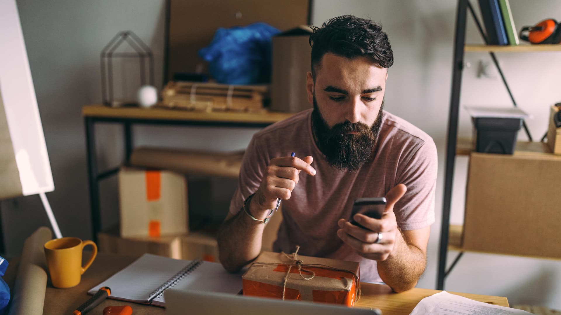 Junger Mann mit Smartphone in der Hand. Vor ihm liegt ein Paket auf dem Schreibtisch.