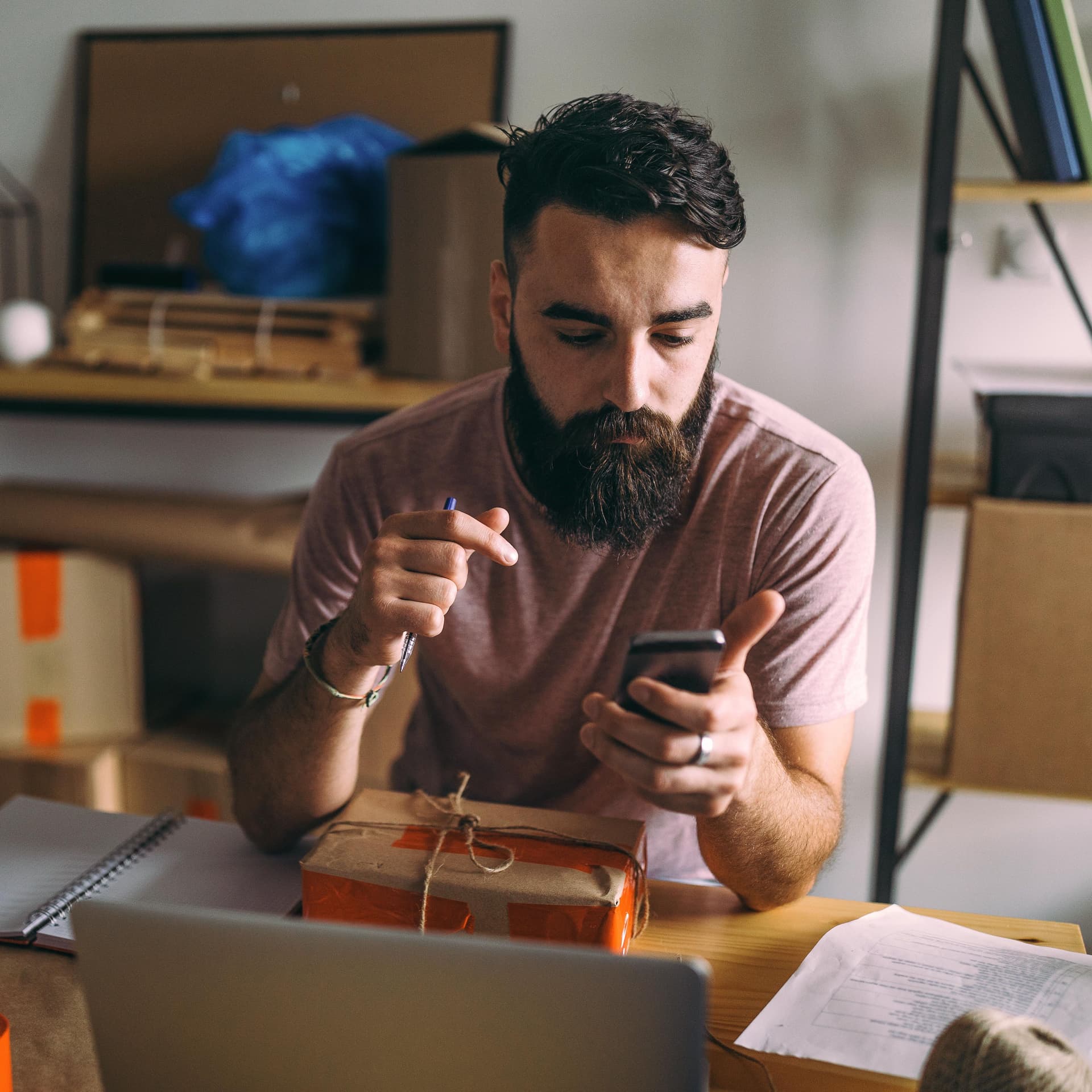 Junger Mann mit Smartphone in der Hand. Vor ihm liegt ein Paket auf dem Schreibtisch.