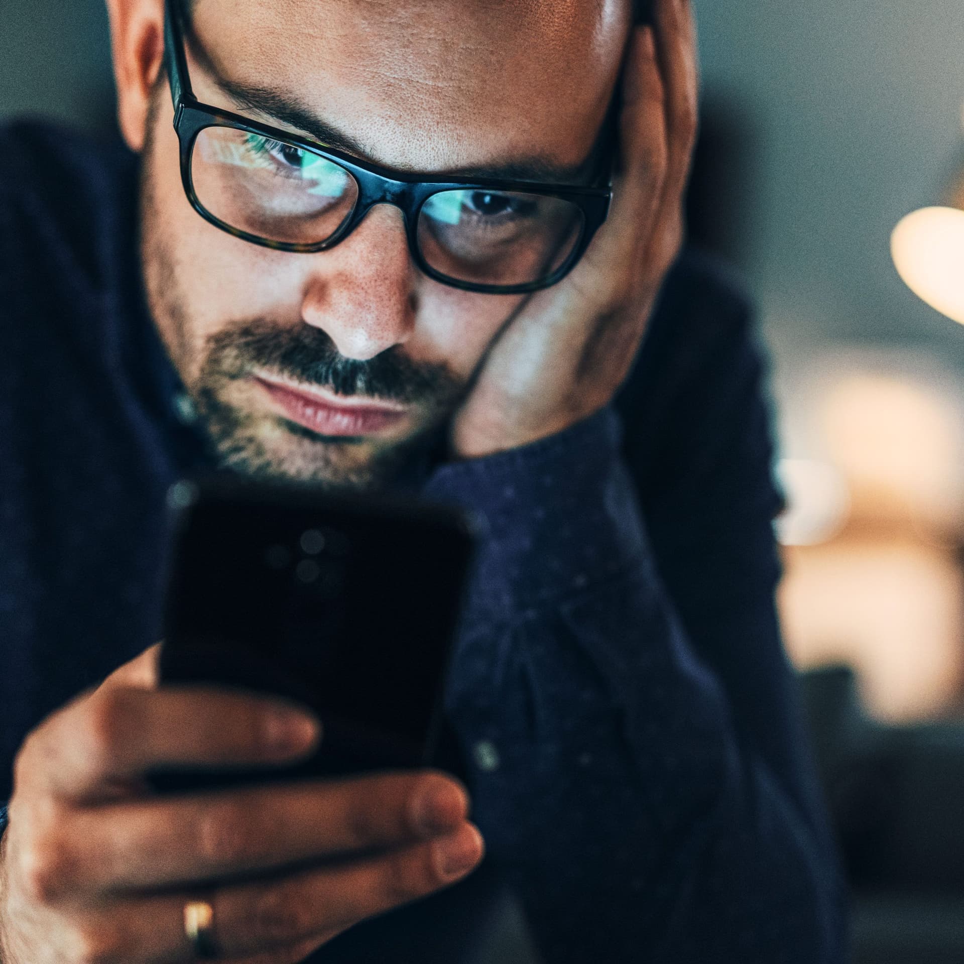 Porträt eines Mannes mittleren Alters. Er hat seinen Kopf aufgestützt und schaut missmutig auf sein Smartphone.