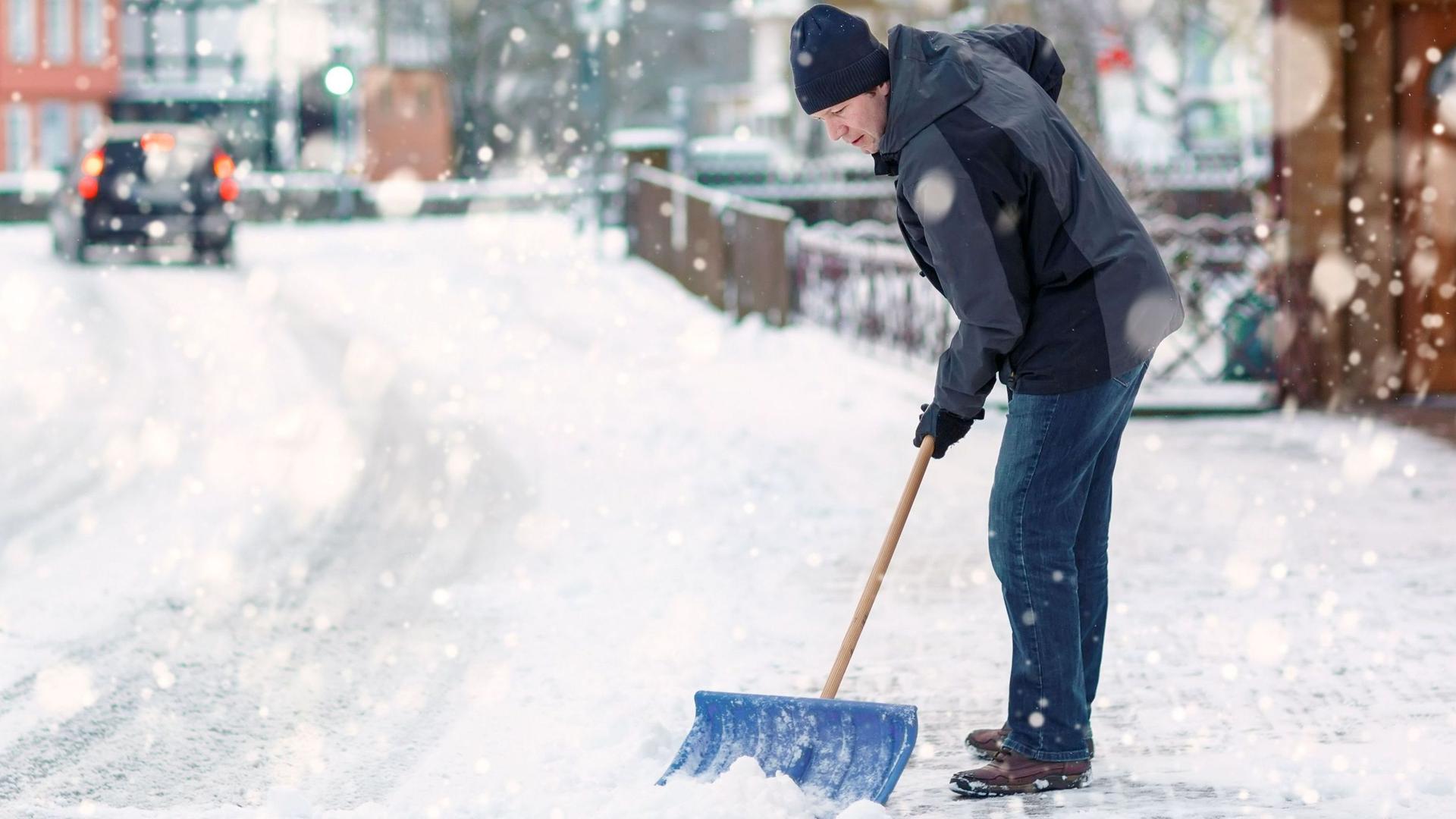 Mann mit blauer Schneeschaufel reinigt Bürgersteig einer ländlichen Straße im Winter.