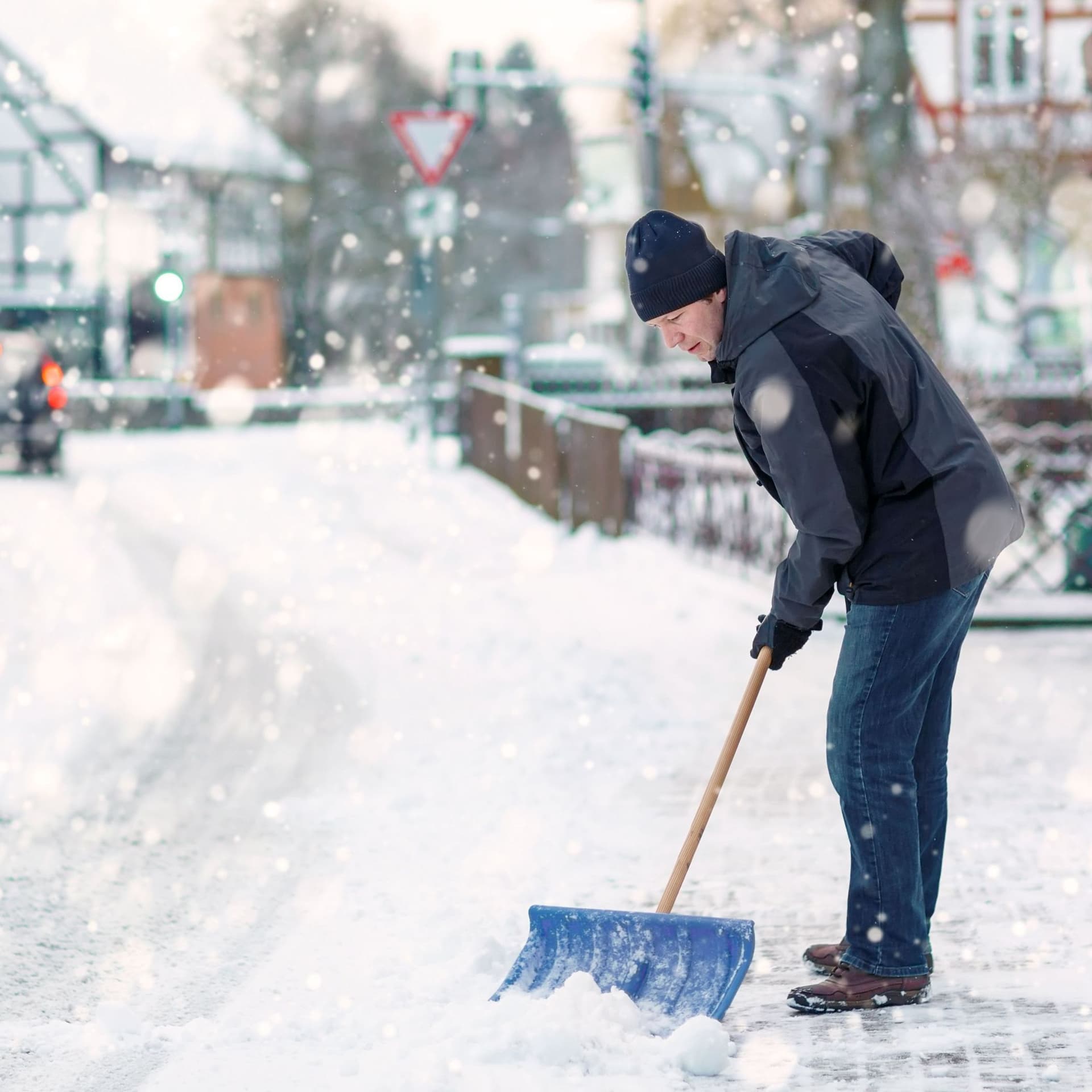 Mann mit blauer Schneeschaufel reinigt Bürgersteig einer ländlichen Straße im Winter.