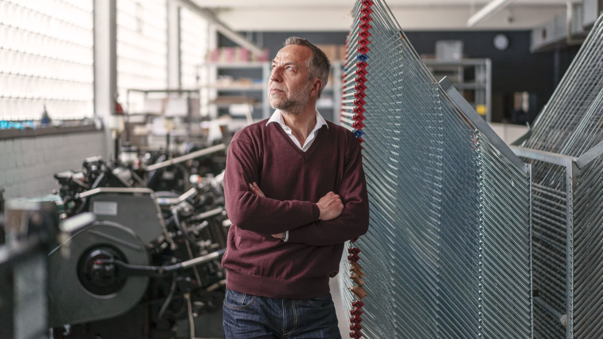 Älterer Mann in einer Druckerei. Er lehnt mit verschränkten Armen an einer Maschine und schaut aus dem Fenster.
