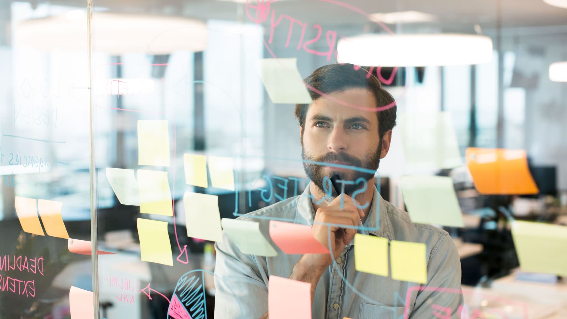 Junger Mann steht vor Glasscheibe die mit Post-Its beklebt und beschriftet ist und schaut konzentriert.