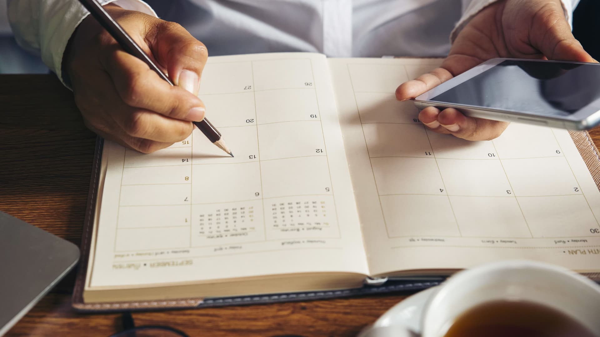 Mann schreibt mit einem Bleistift in einen Kalender. Er hält ein Smartphone in der linken Hand.