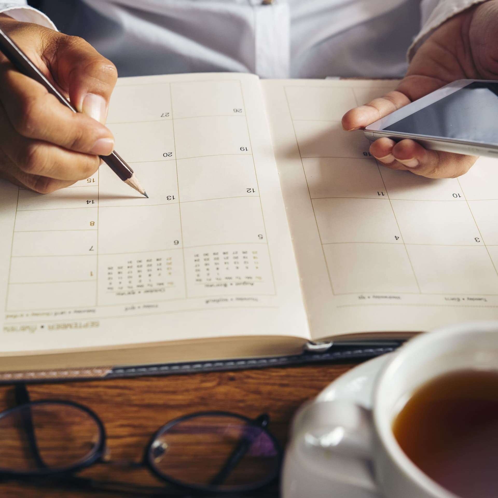 Mann schreibt mit einem Bleistift in einen Kalender. Er hält ein Smartphone in der linken Hand.
